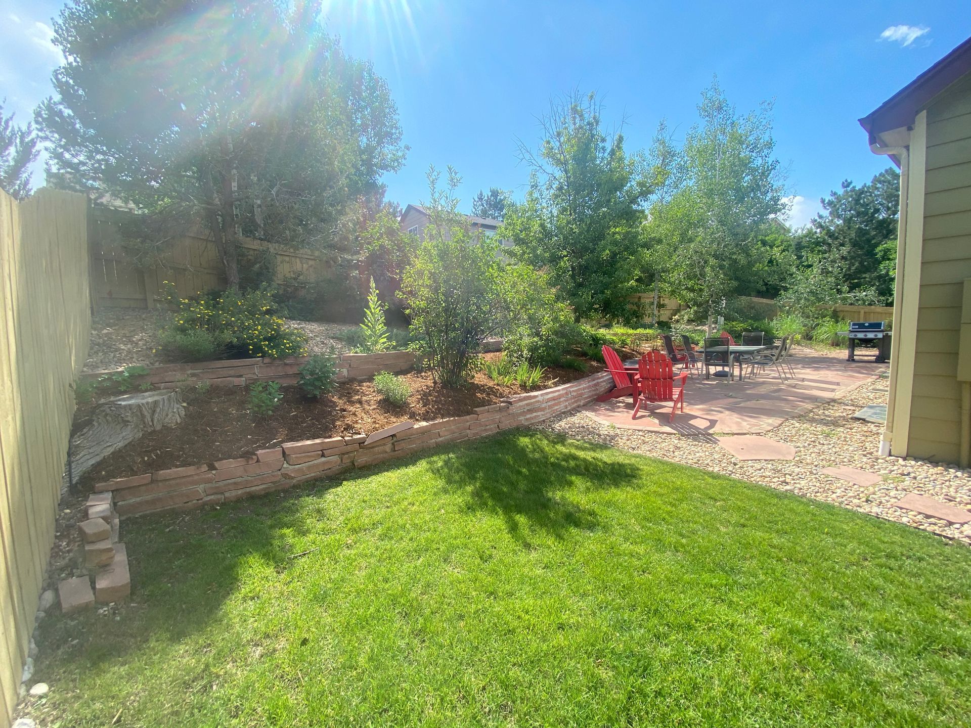 Backyard with tiered landscaping, lawn, patio with red chairs, and blue sky.