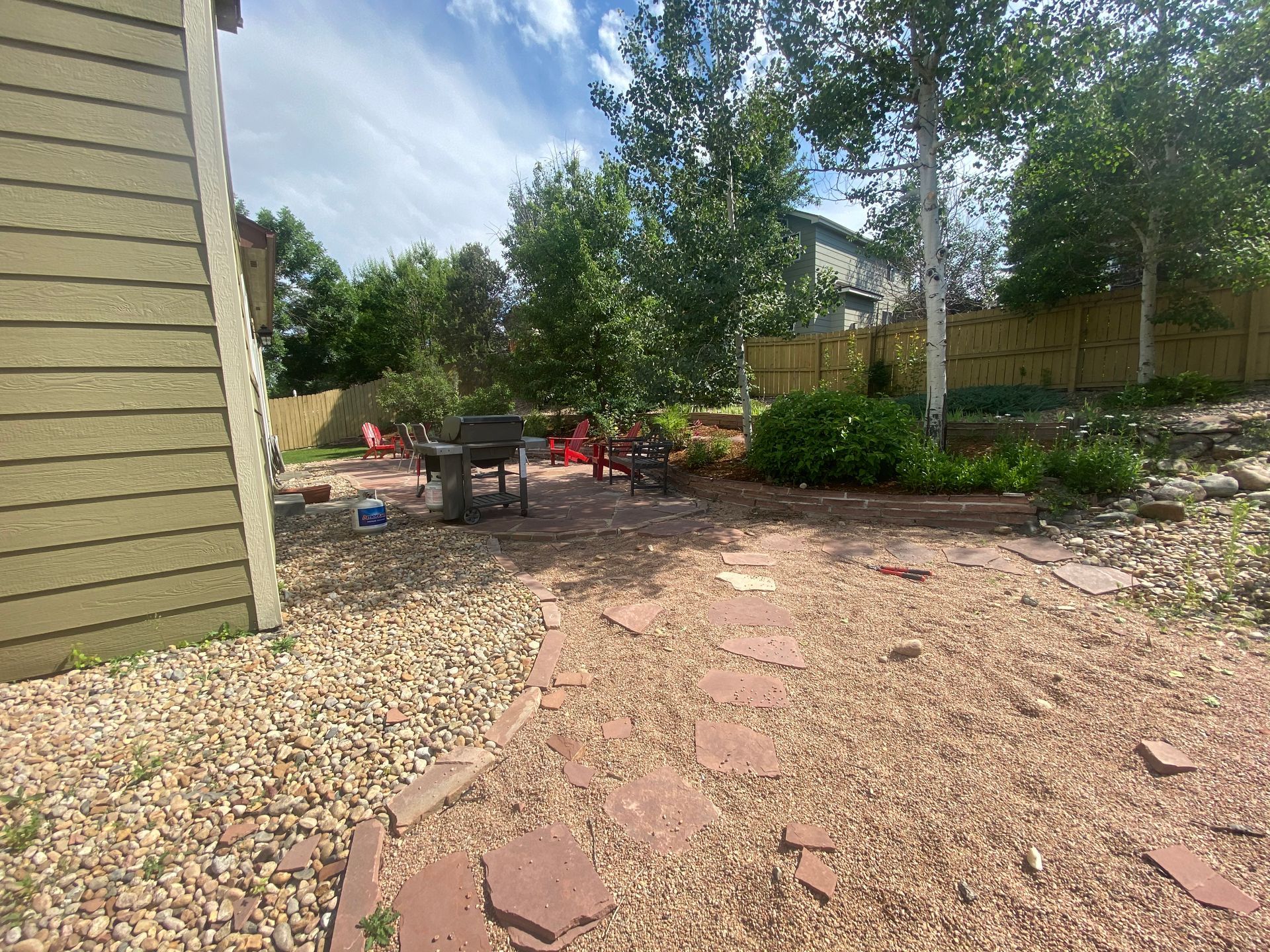 Backyard with a gravel and stone patio, grill, trees, and fence on a sunny day.