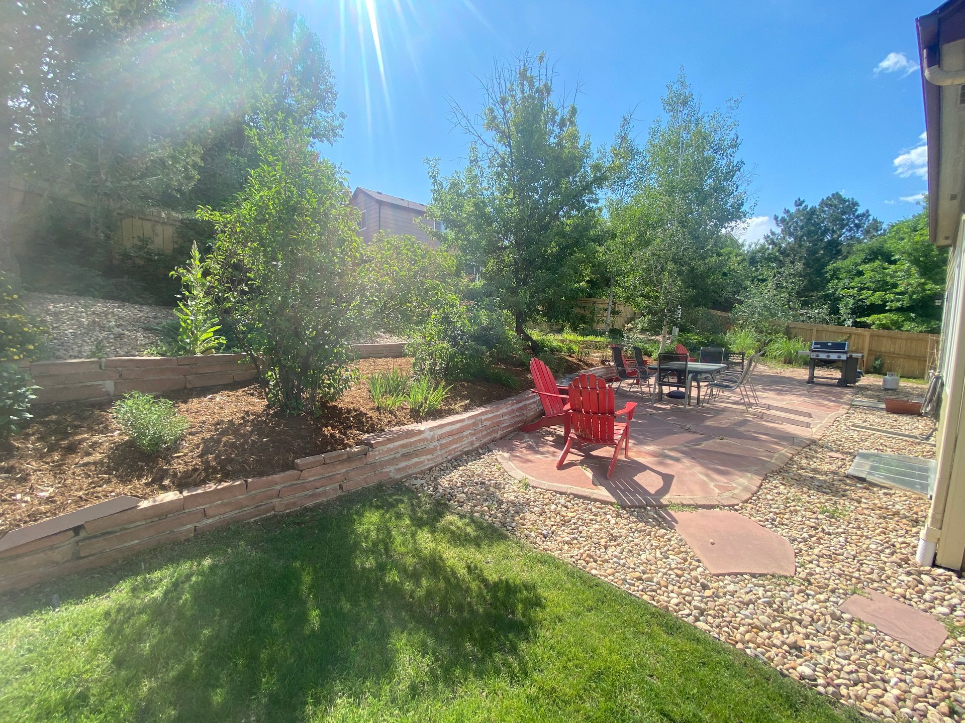 Sunny backyard with red chairs, a patio, a grill, and tiered landscaping.
