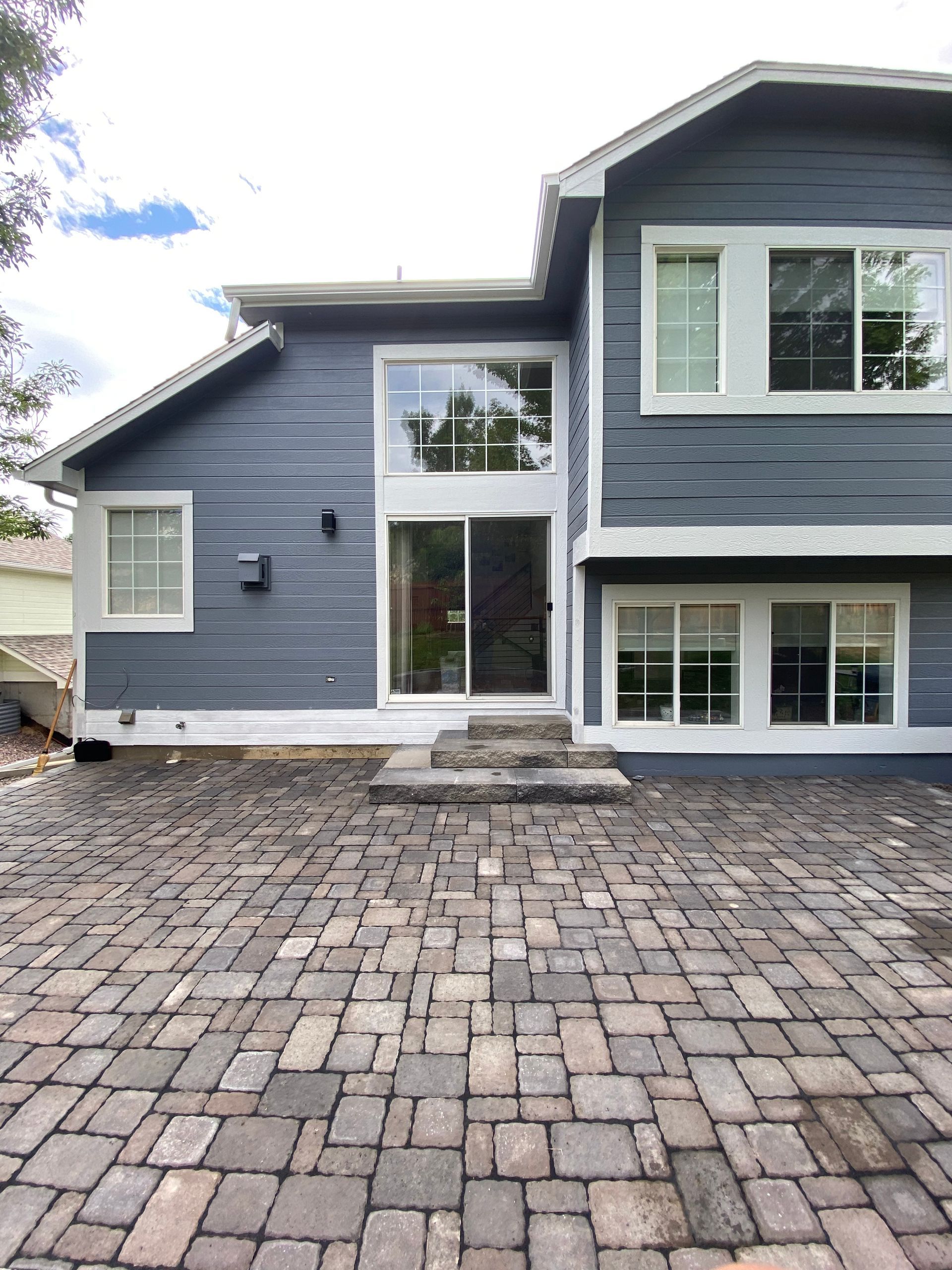 Two-story house with dark blue siding, white trim, and a brick patio. A sliding glass door and large windows are visible.