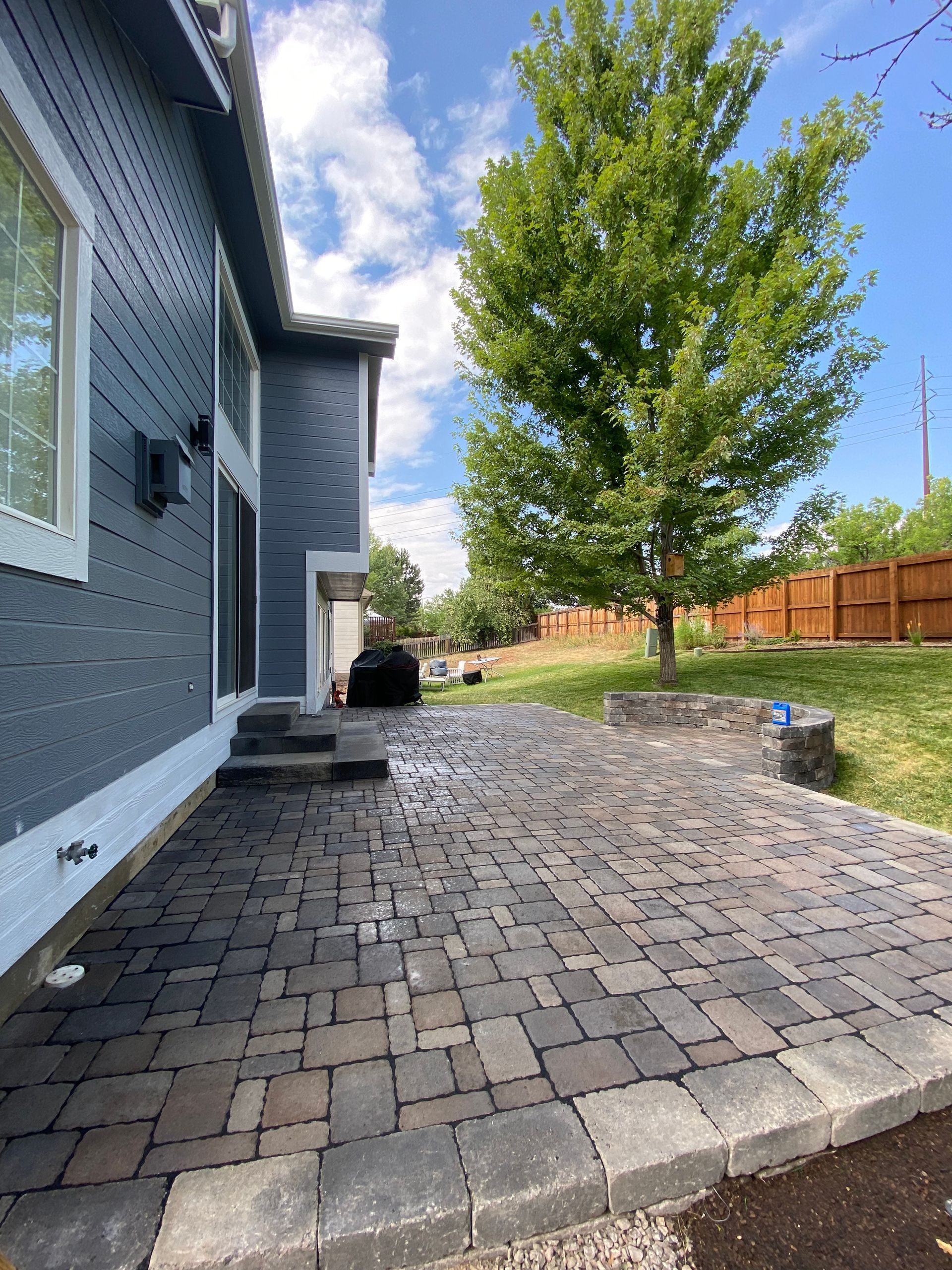 Backyard patio with brick pavers next to a gray house, tree, and green grass under a blue sky.