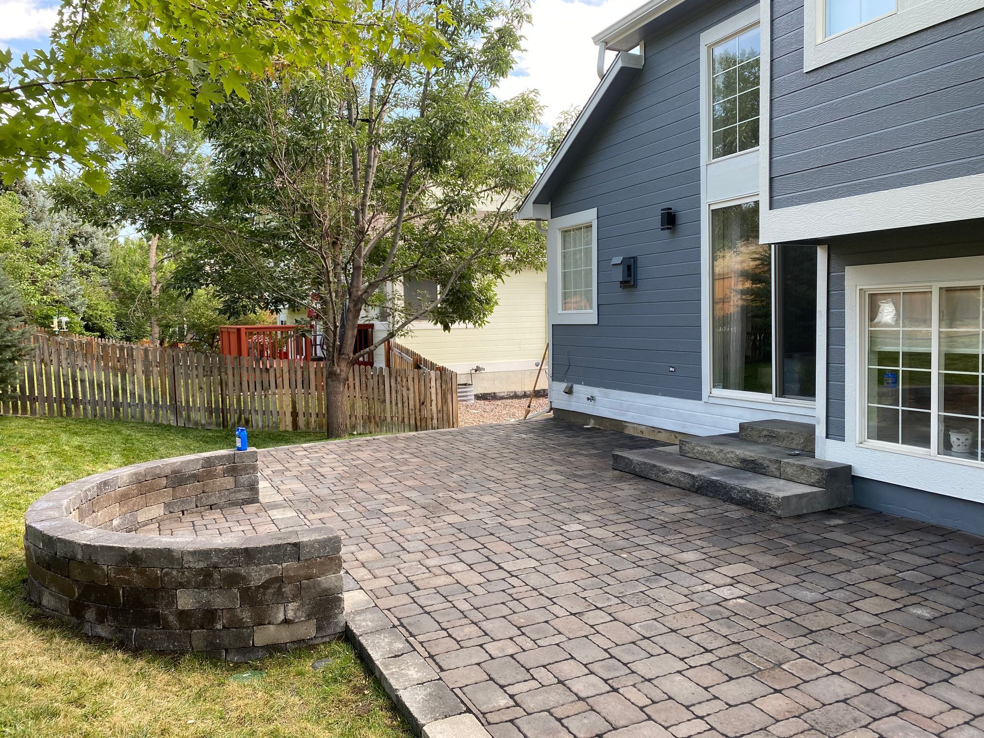 A brick patio with a low curved wall next to a blue house with steps and windows. Green grass and trees surround it.