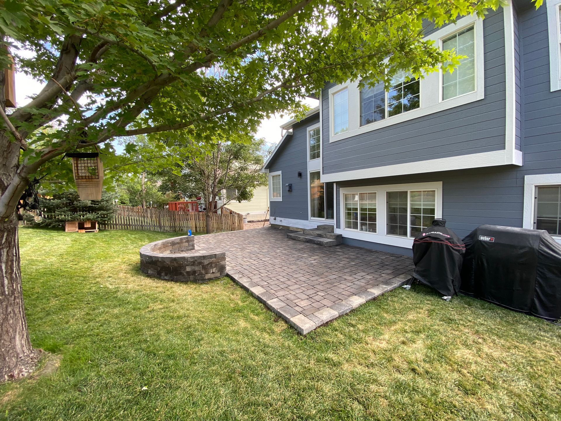 Backyard patio with fire pit, grill, and gray house under a large tree.