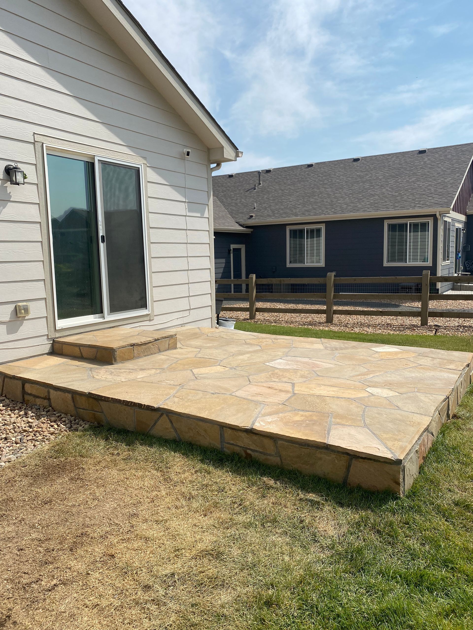 Stone patio outside a white house with a sliding glass door. A blue house with a wooden fence is in the background.