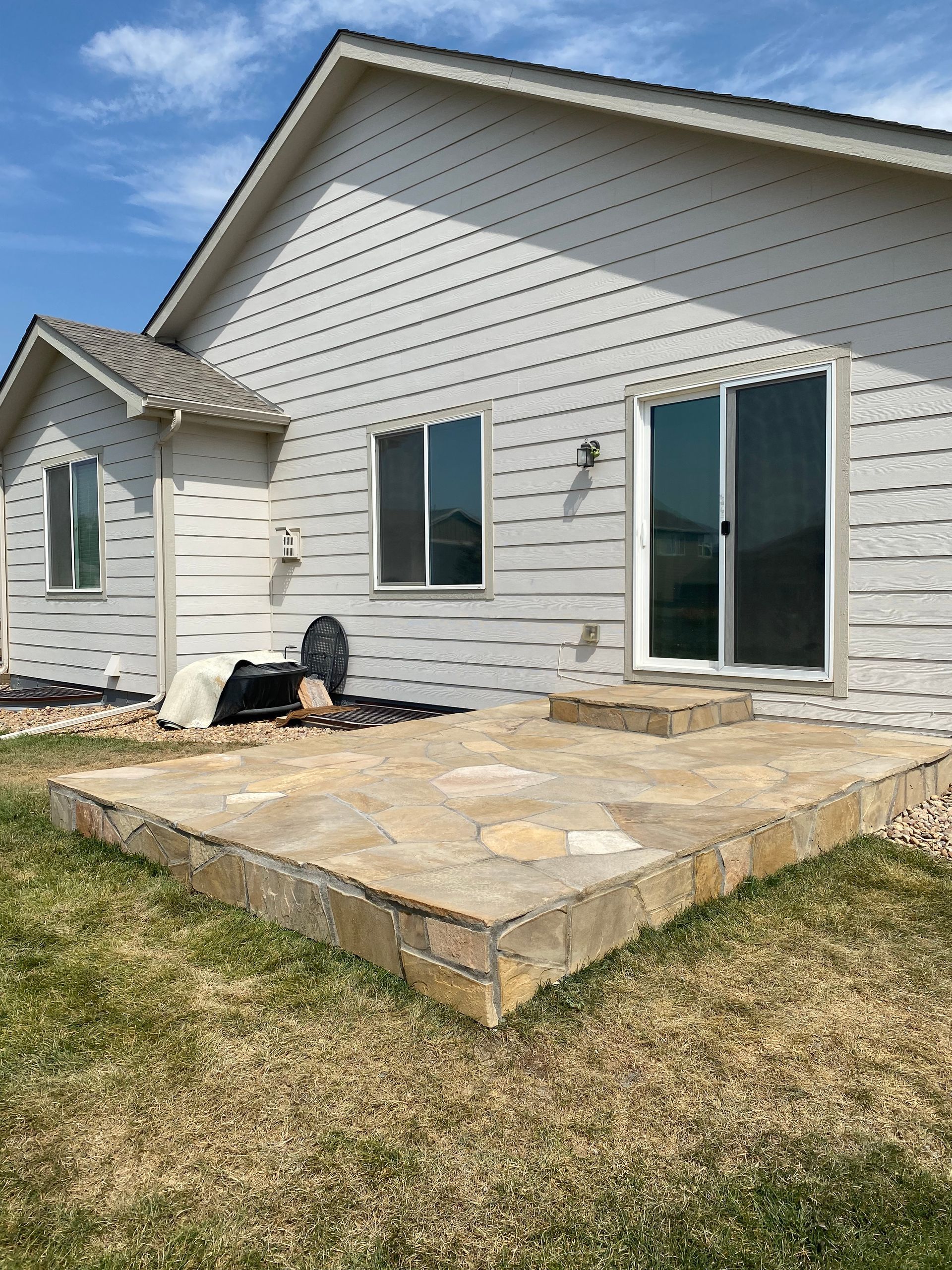 Backyard patio with light tan pavers, adjacent to a light-colored house with a sliding glass door.