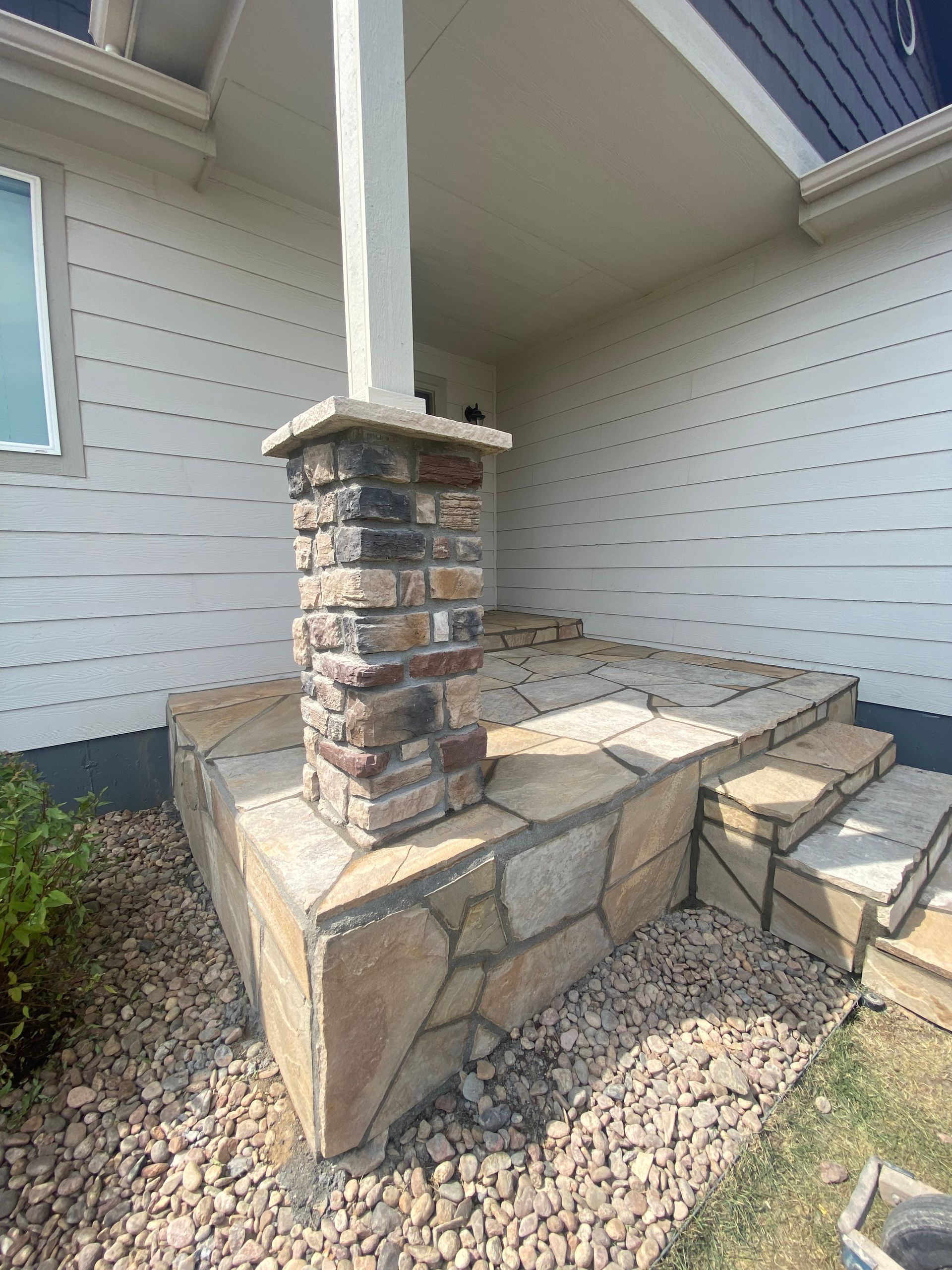 Stone porch with steps and column, beige siding, gravel ground.