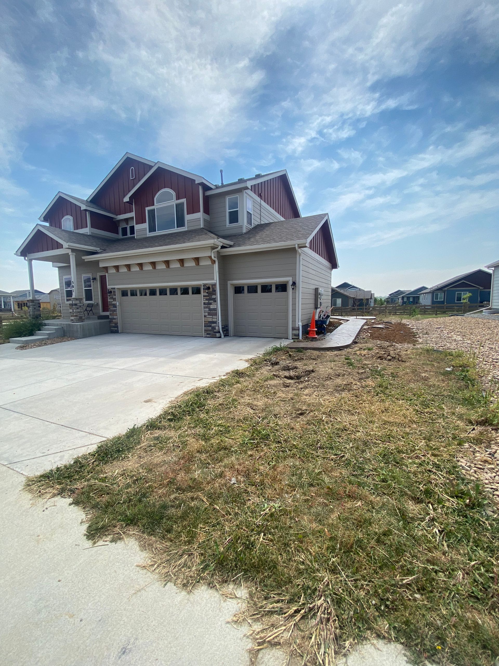Two-story house with red roof trim, beige siding, three-car garage, concrete driveway, and grassy foreground under a blue sky.