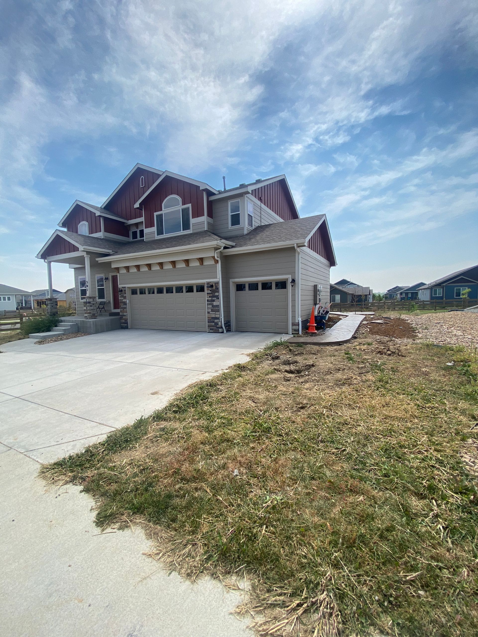 Two-story house with a tan exterior, red trim, and three-car garage. Driveway and unkempt lawn in the foreground. Blue sky.