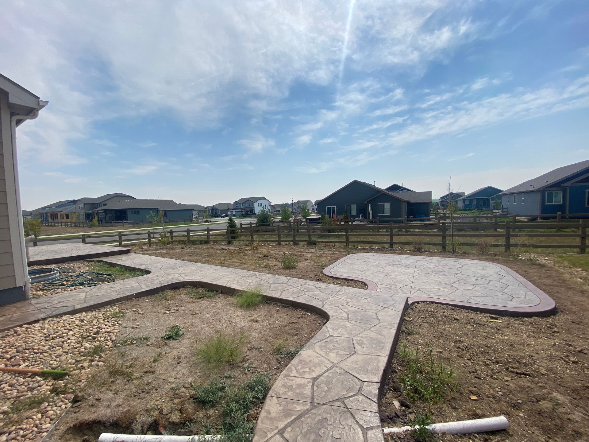 Backyard with stamped concrete patio and path, brown yard, houses and fence in the distance, sunny blue sky.