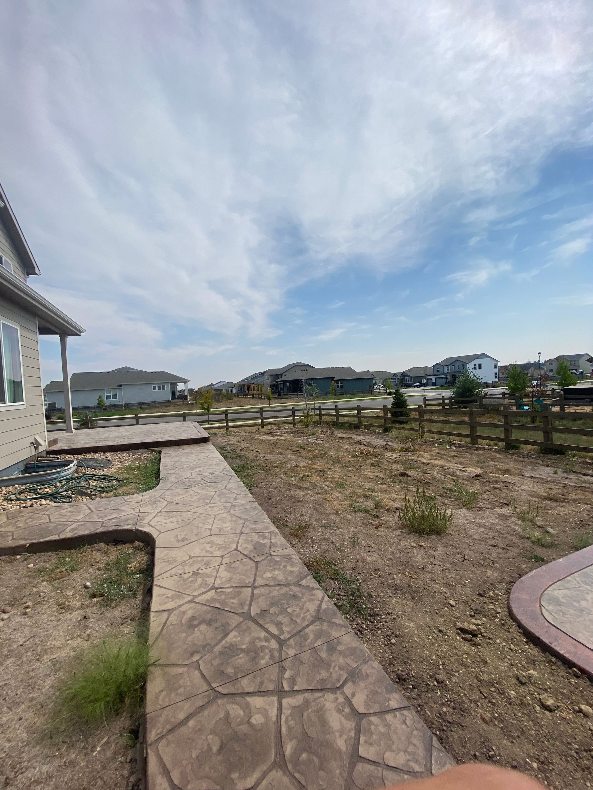 Backyard view: stamped concrete path, dry grass, houses in the distance, blue sky with clouds.