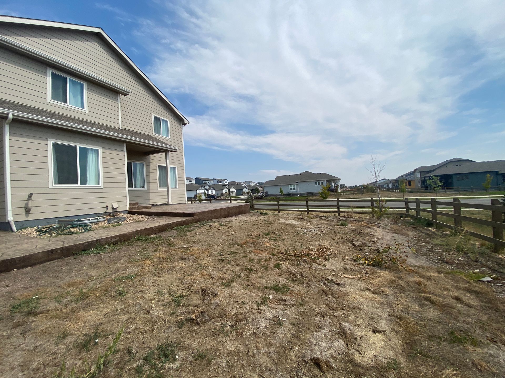 Backyard view of a two-story beige house with dry grass, wooden fence, and blue sky.