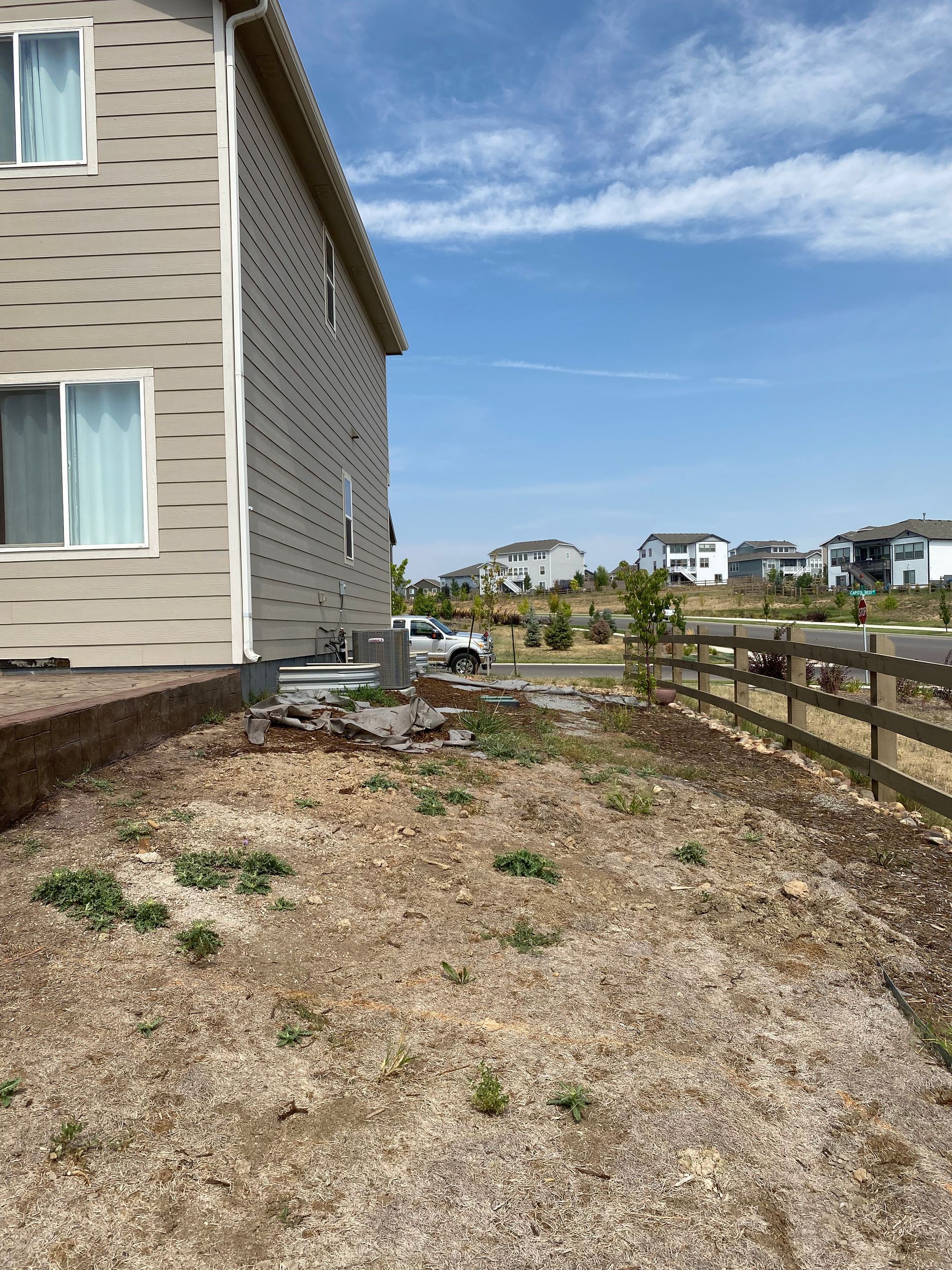 Side of a two-story beige house next to a gravel yard with a fence and a distant neighborhood under a blue sky.