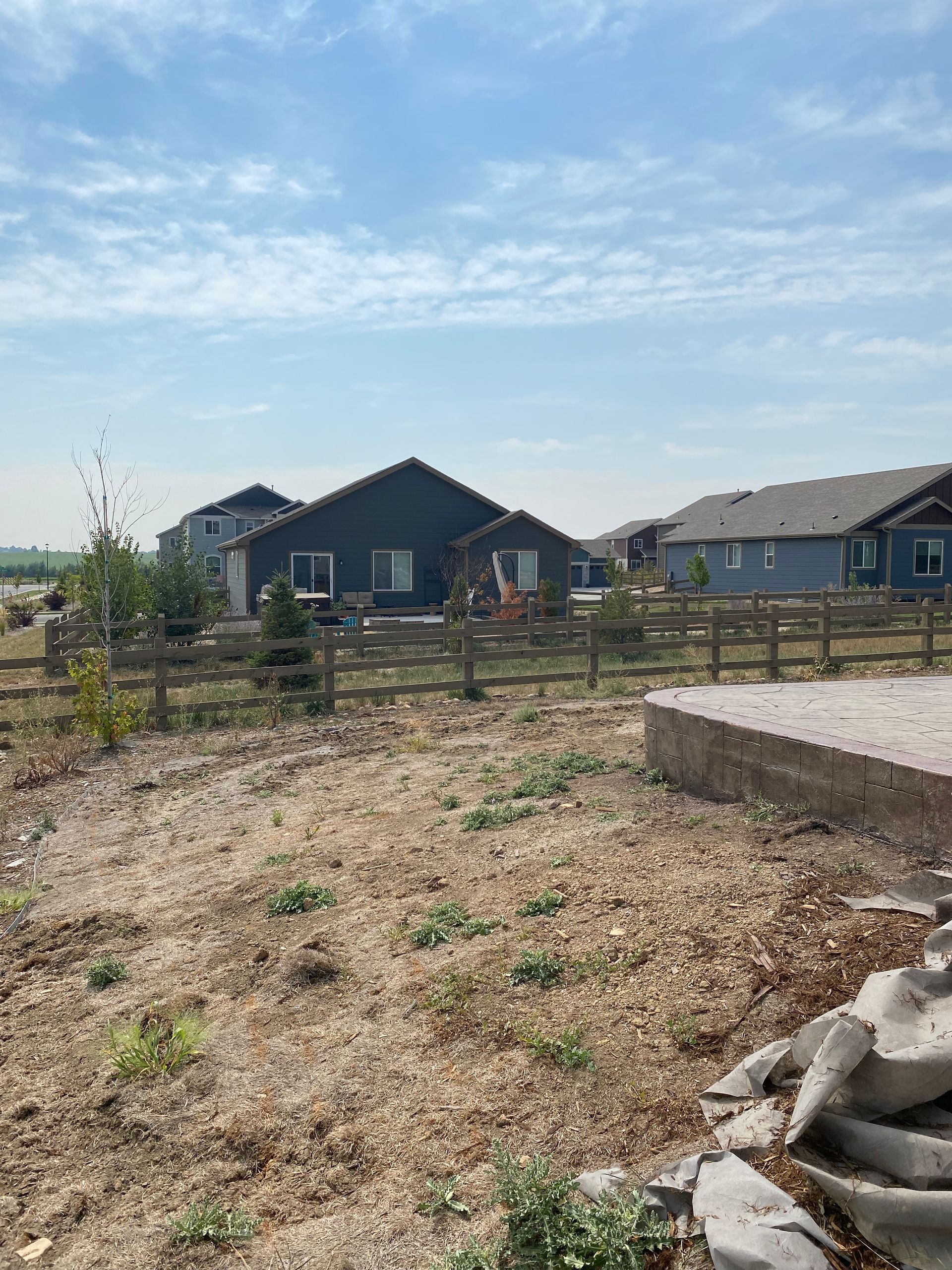 Backyard view with a dark house, wooden fence, and gravel ground under a blue sky.