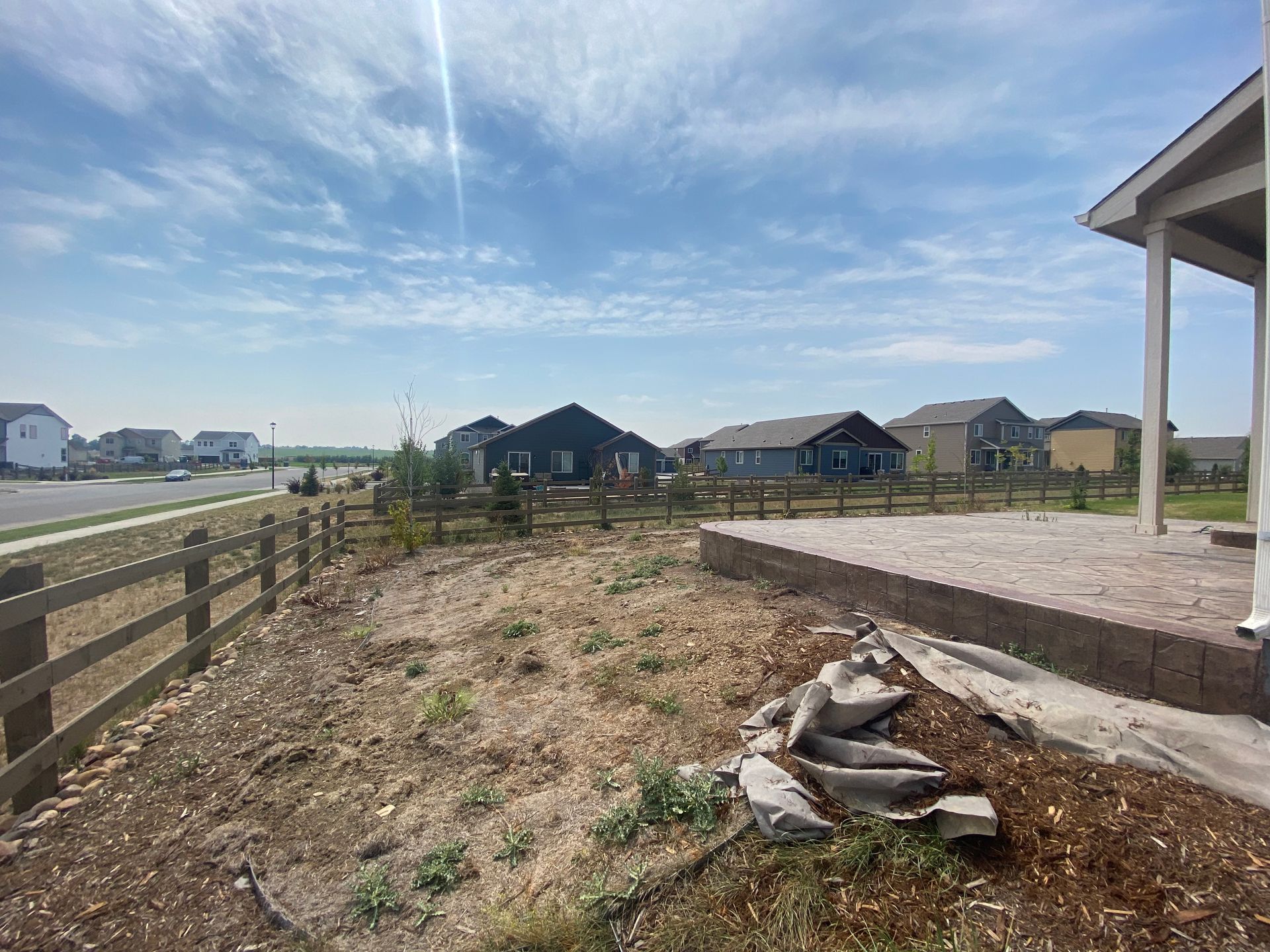 Backyard with a brown fence, dried grass, houses, and a blue sky with sunlight.