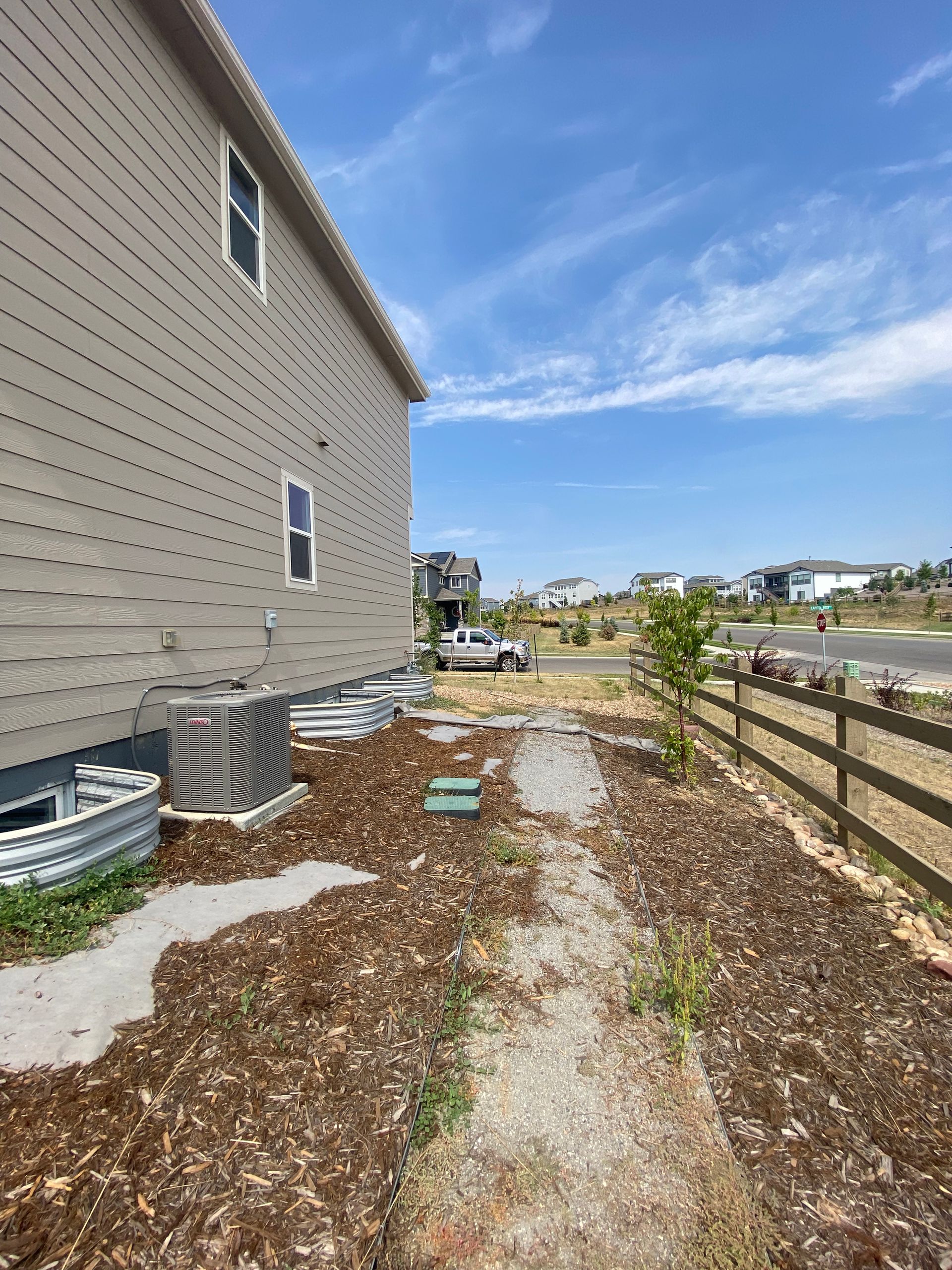 Side of a house with a pathway, mulch, and fence under a blue sky.