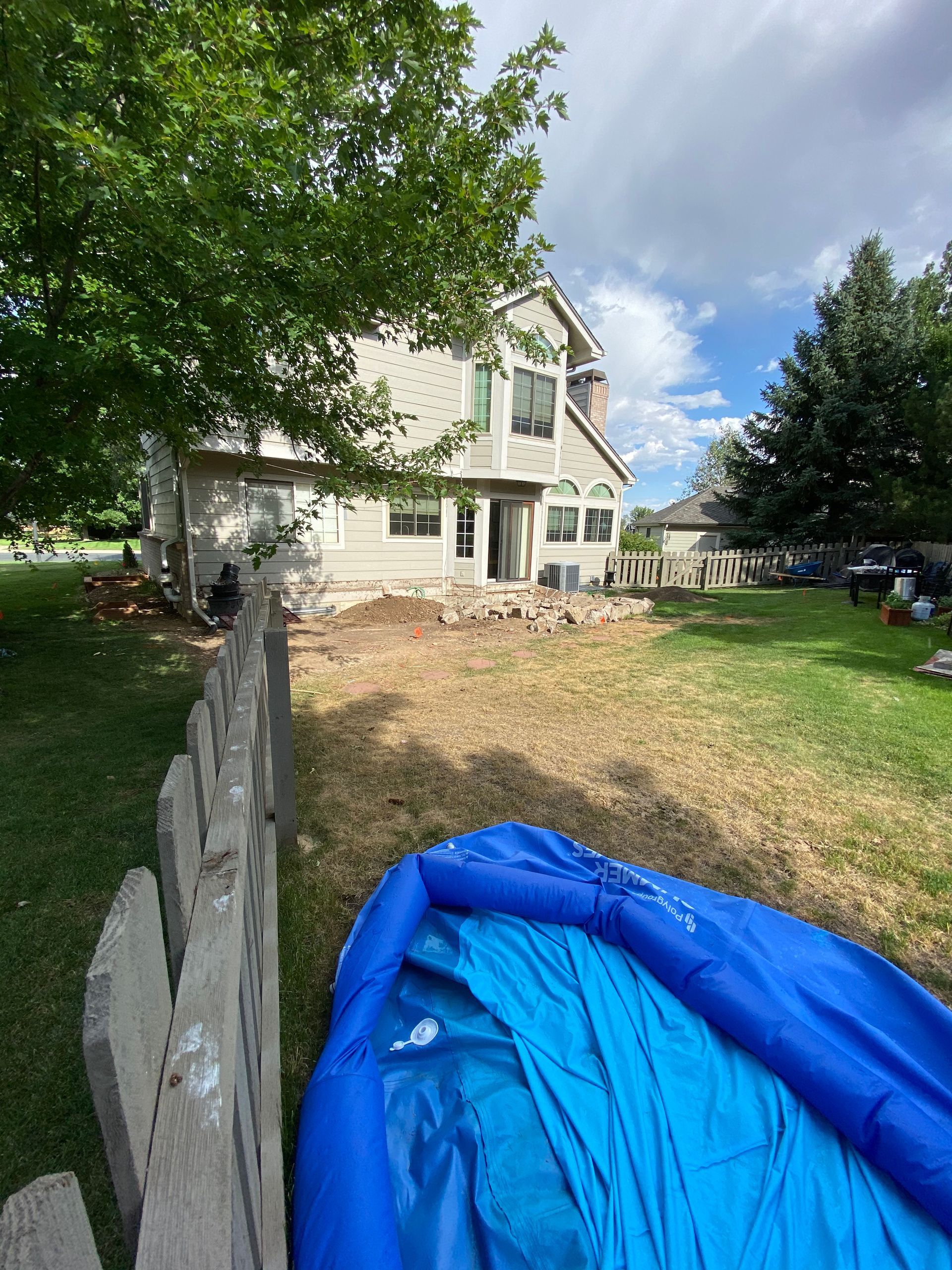 Backyard view with a house, a fence, and a blue inflatable pool on the dry, patchy lawn under a cloudy sky.