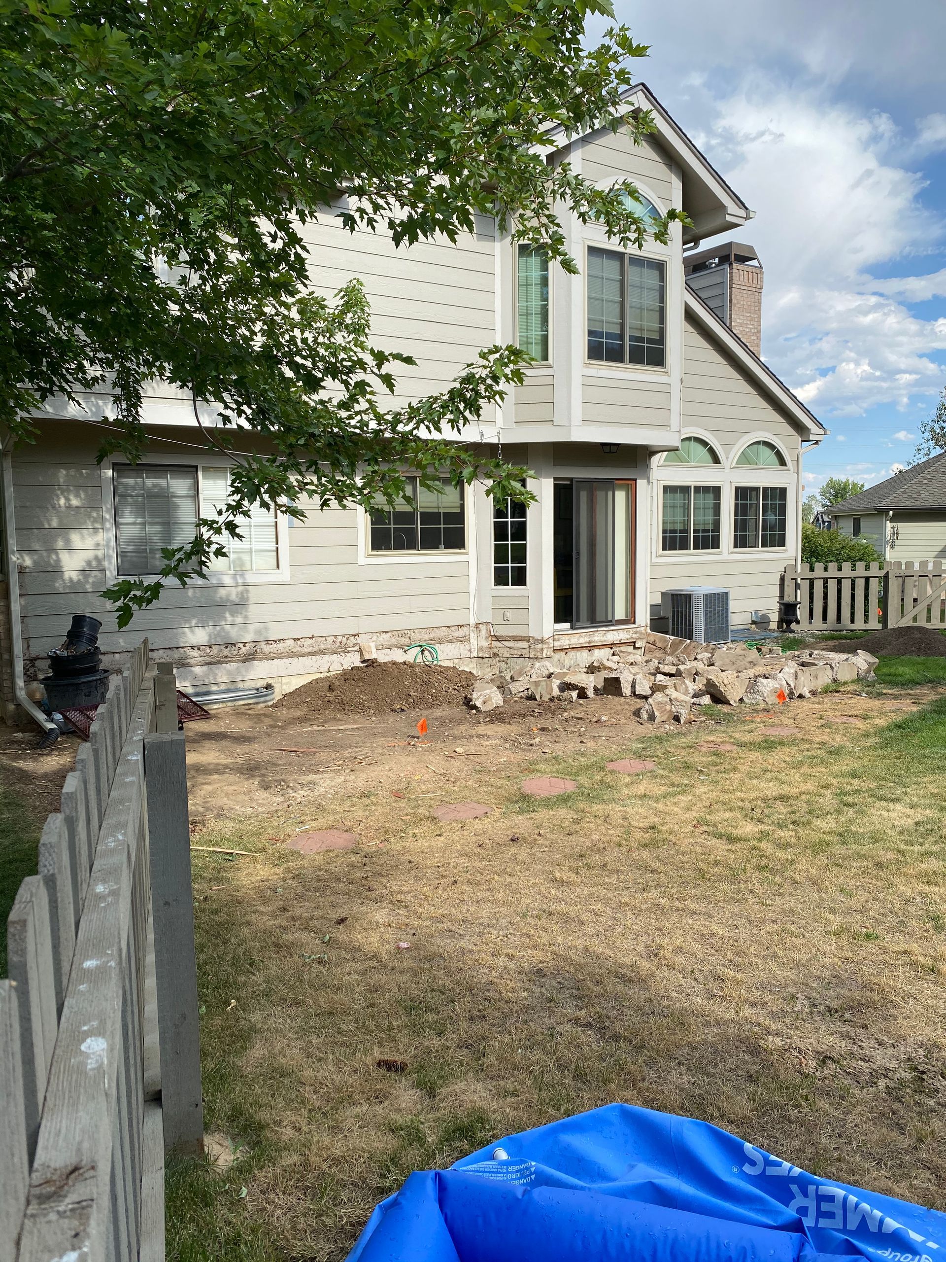 Backyard with house under construction, exposed foundation, and pile of bricks. Dry grass and a blue tarp.
