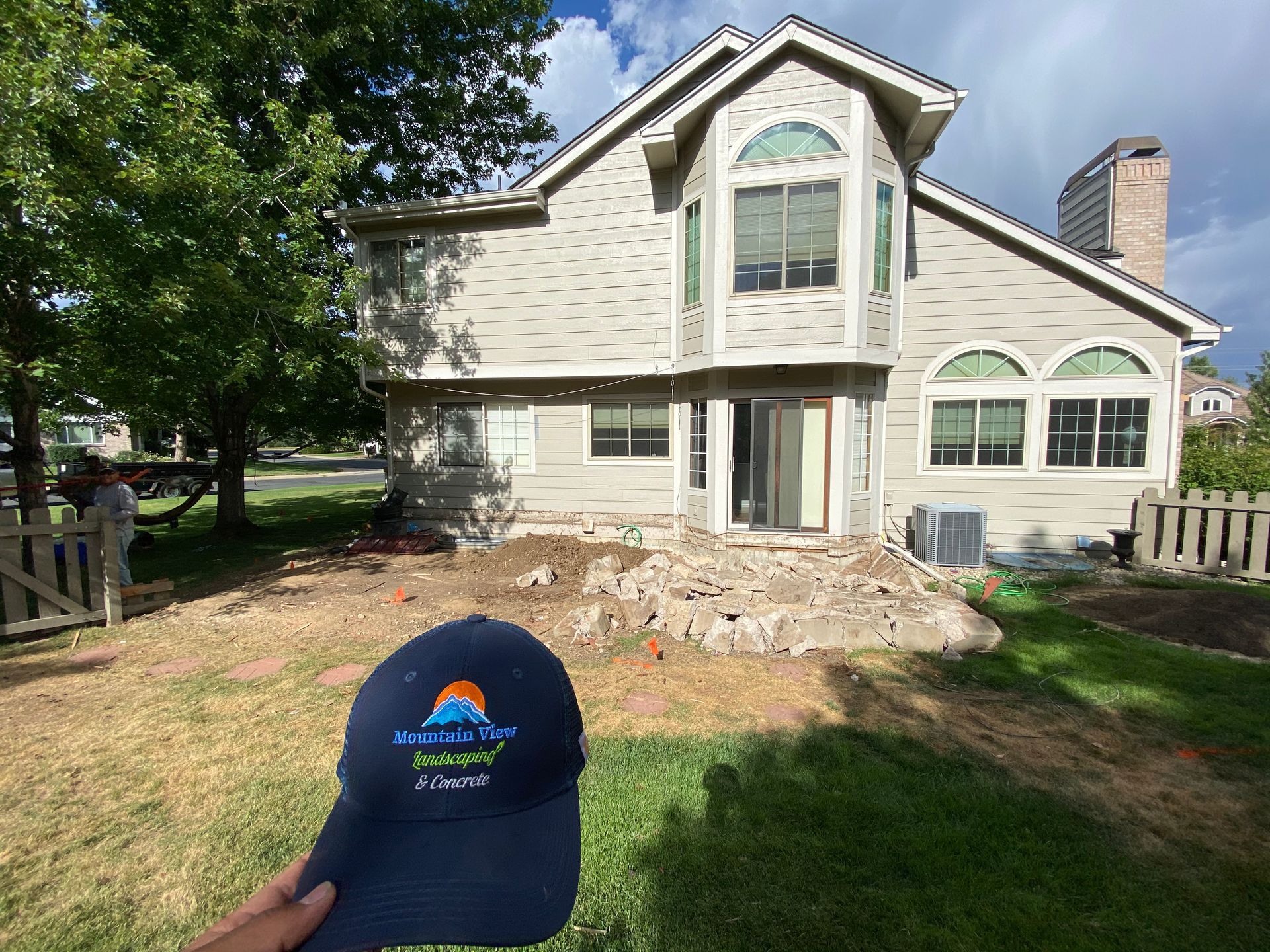 Backyard construction site with a house, pile of dirt, fence, and a baseball cap held up in the foreground.