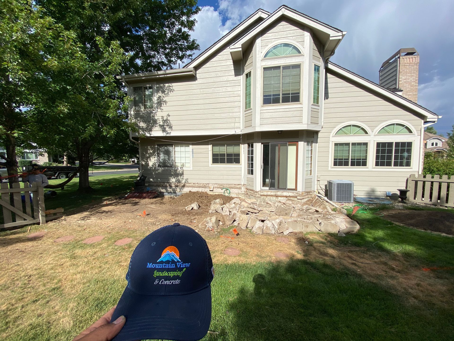 A person holding a hat in front of a house under construction; dirt and debris are in the foreground.