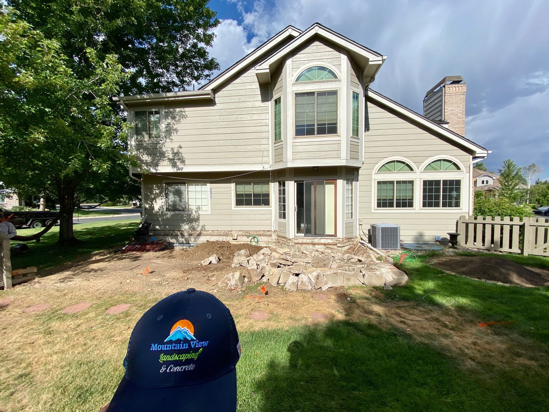 Backyard of a house with construction debris. Tan siding, blue sky, green grass. A person in a hat is in the foreground.