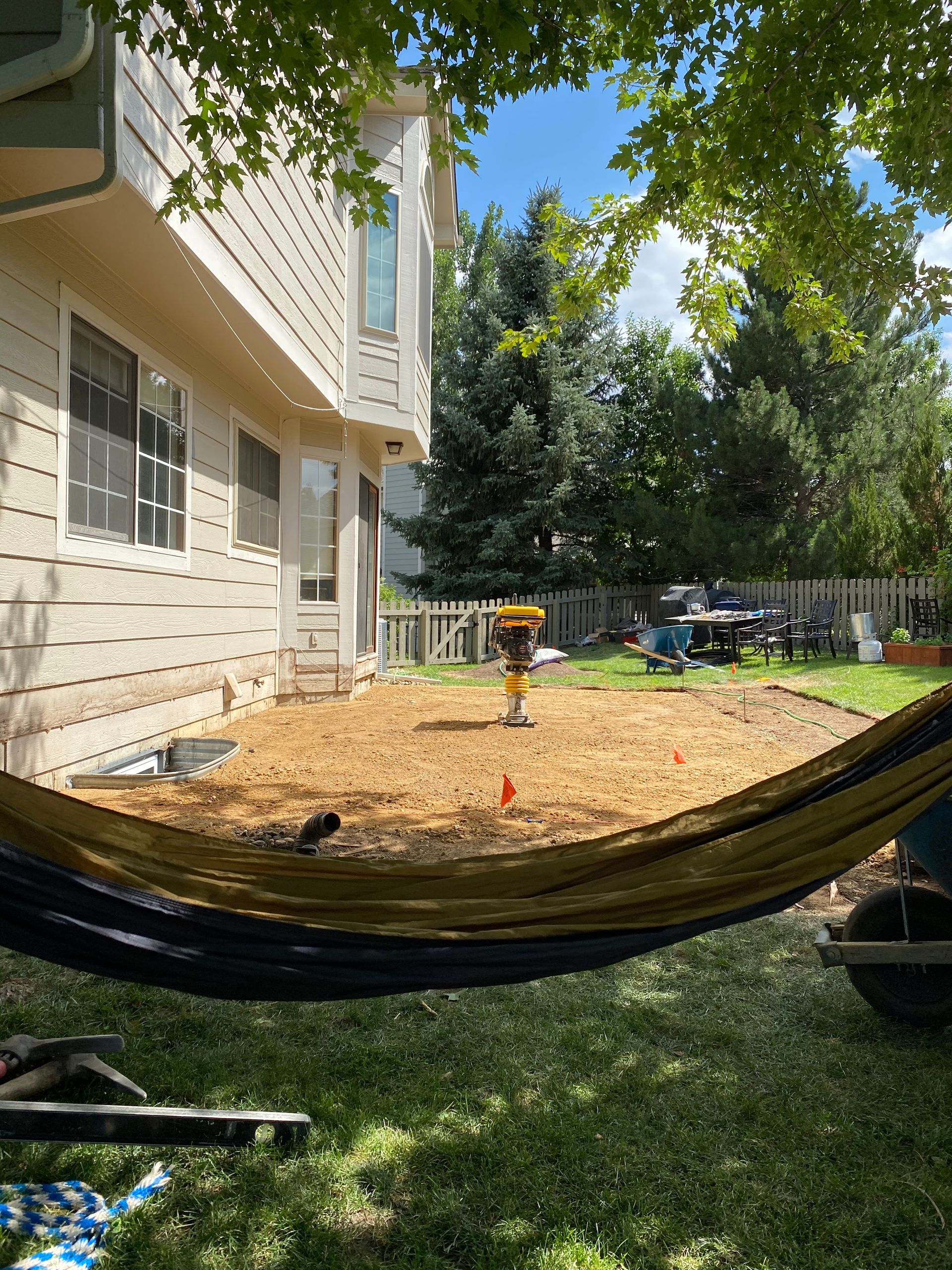 Backyard scene with construction equipment; dirt ground, two-story house, and hammock in the foreground.