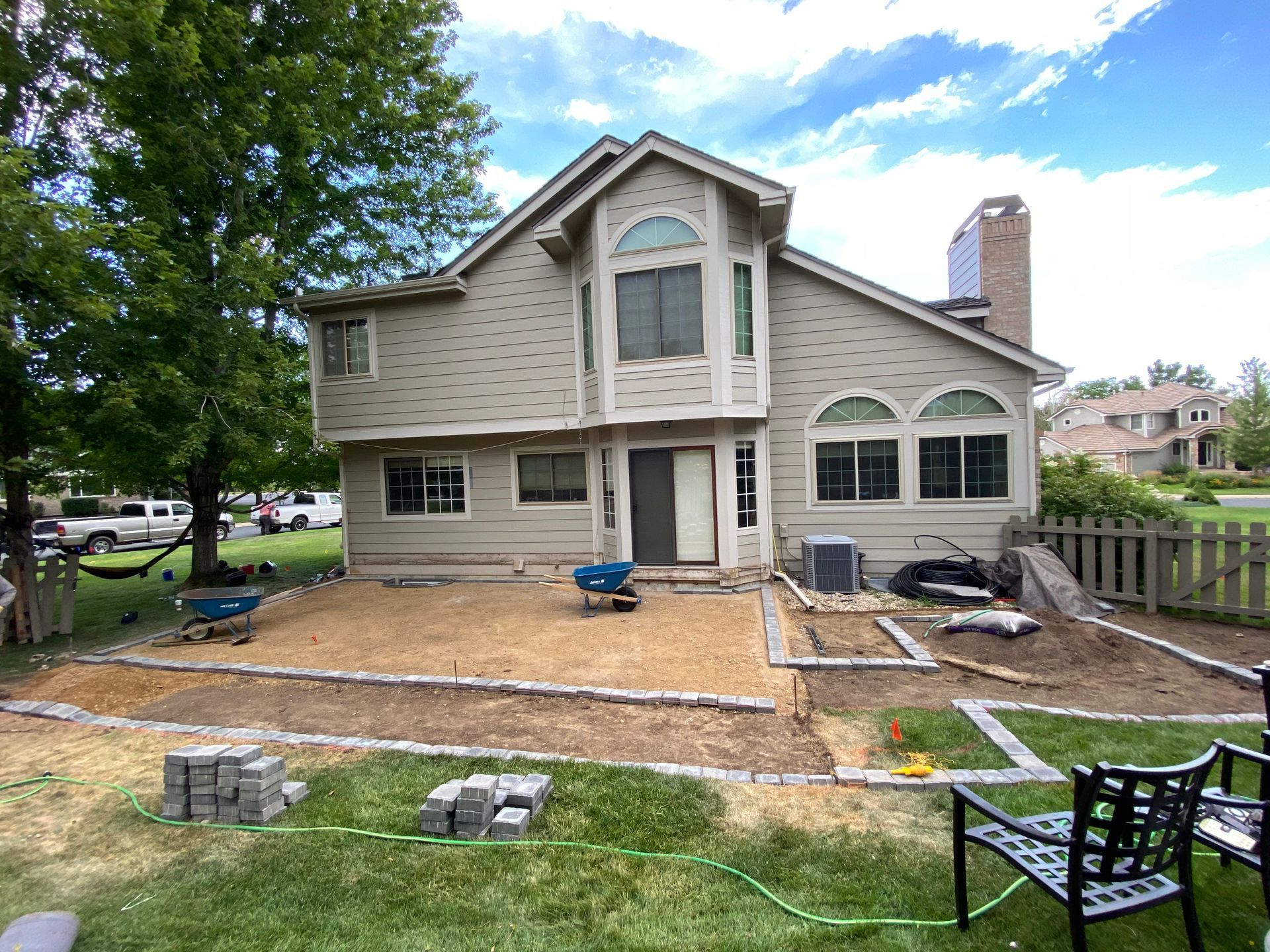 Backyard patio under construction; tan siding on two-story house, paver stones, wheelbarrows, green lawn, blue sky.