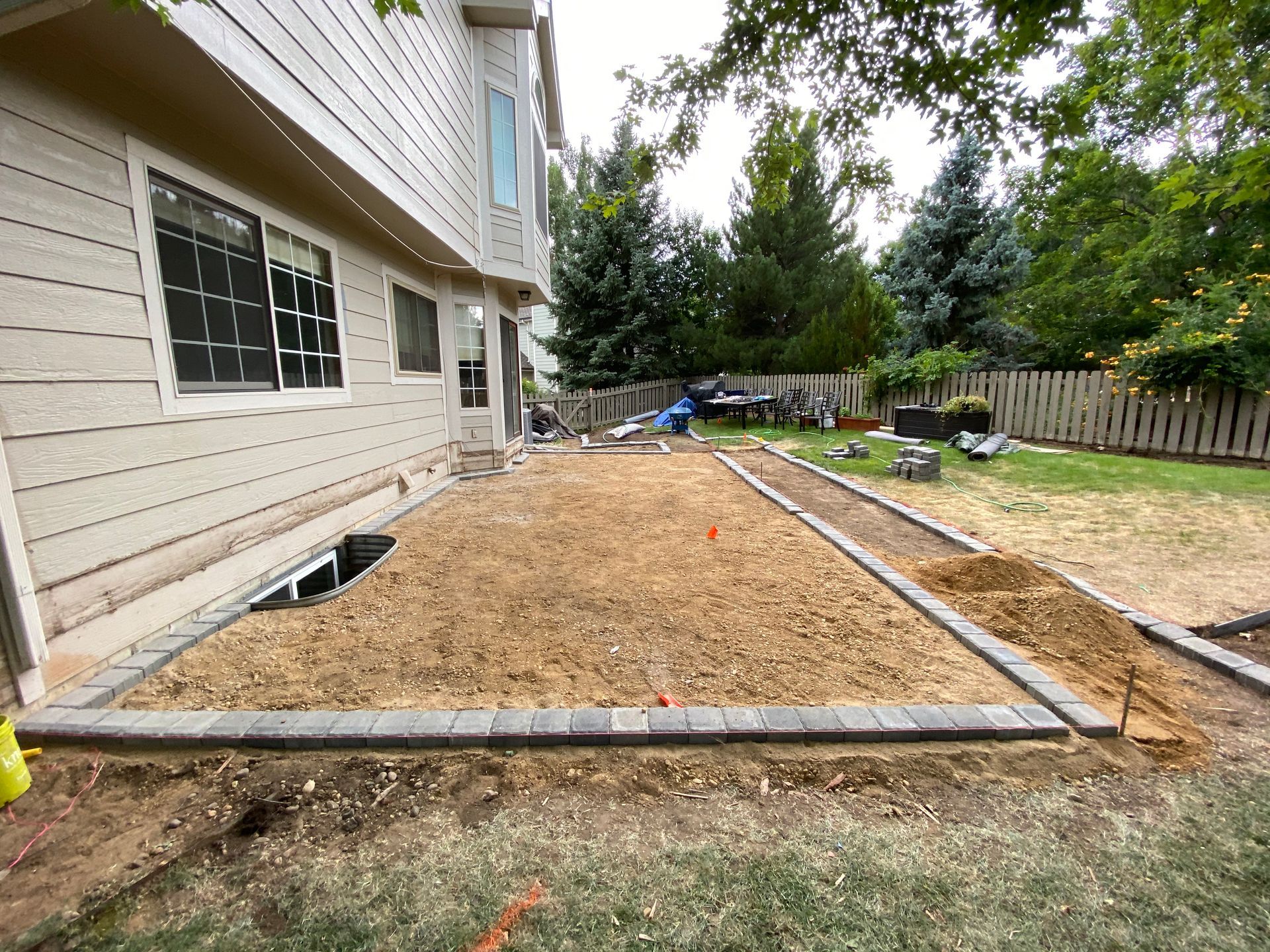 A backyard patio under construction, bordered by gray pavers and a light-colored building.