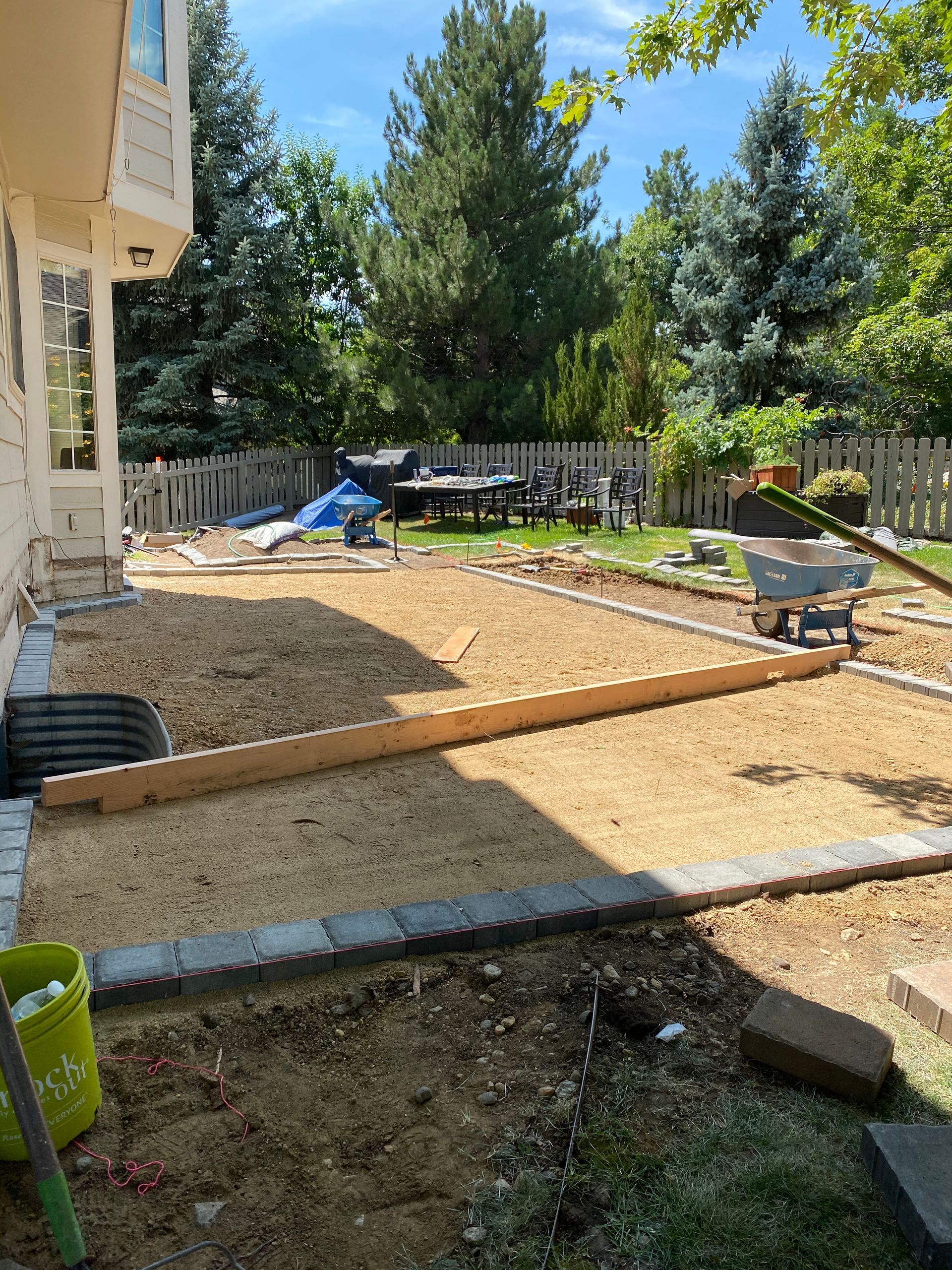 Backyard with construction: gravel base, edging, house on left, trees, play set, and a wheelbarrow.