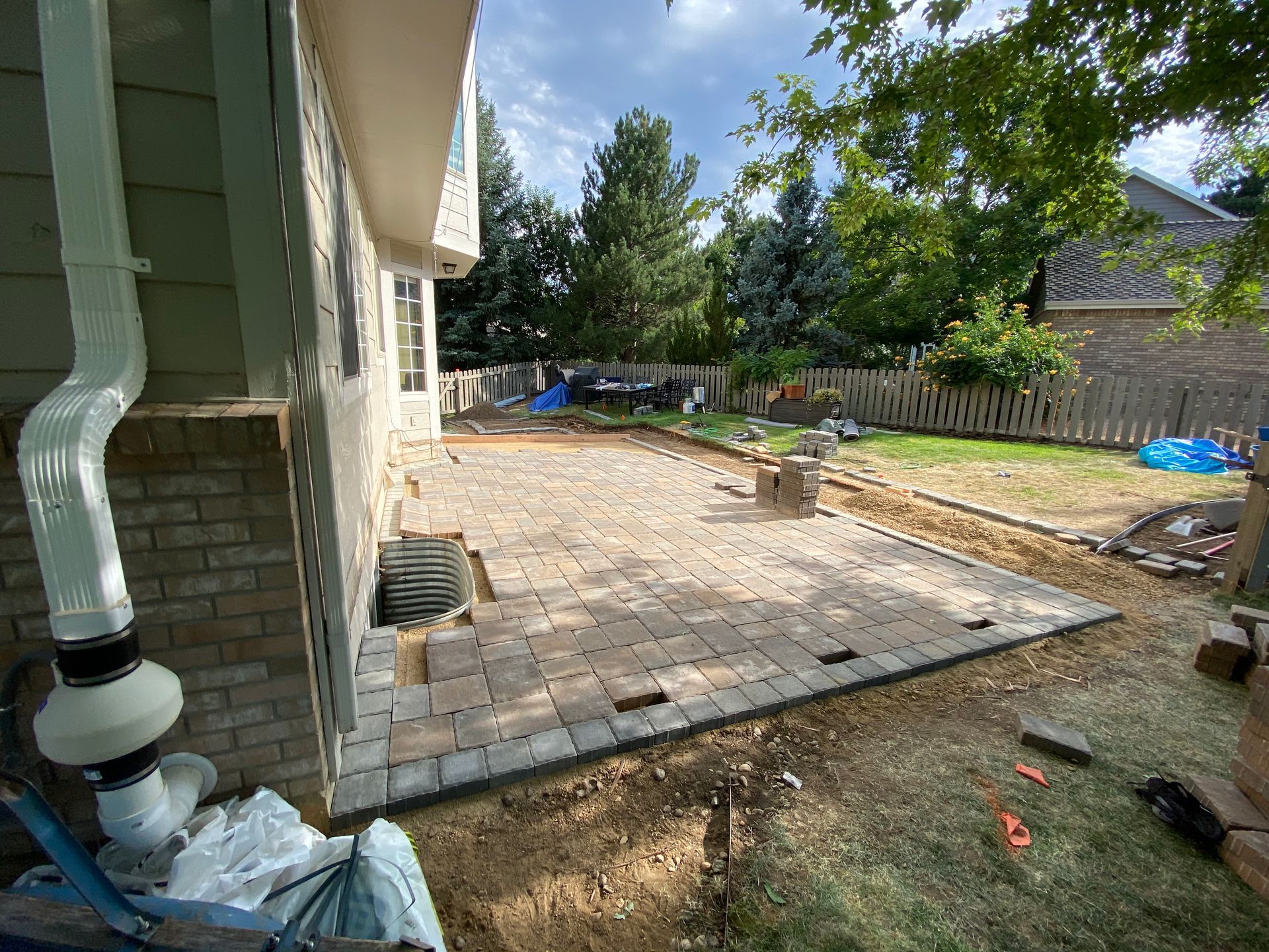 Backyard patio under construction; brick house, pavers laid, dirt, and grassy yard.
