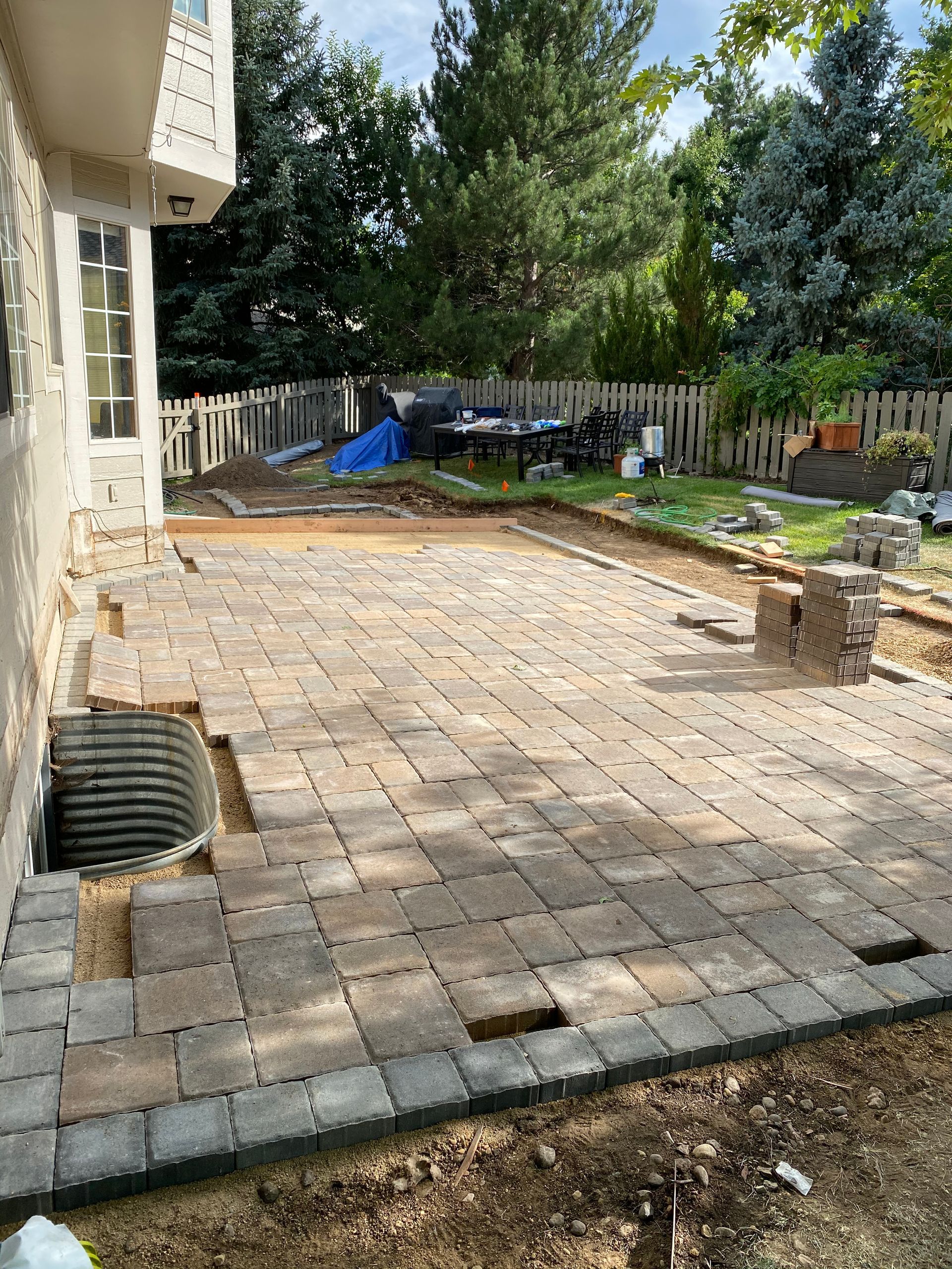 Paver patio under construction next to a house with exposed dirt and construction materials in a yard.