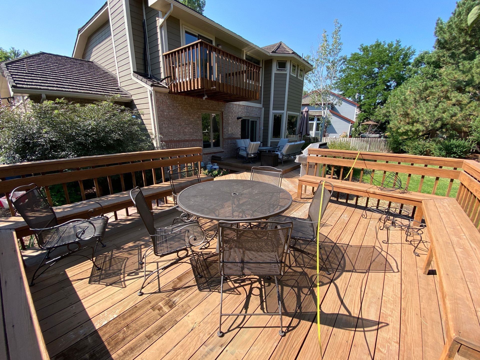 Wooden deck with patio furniture, attached to a multi-level house with a balcony, trees in the background.