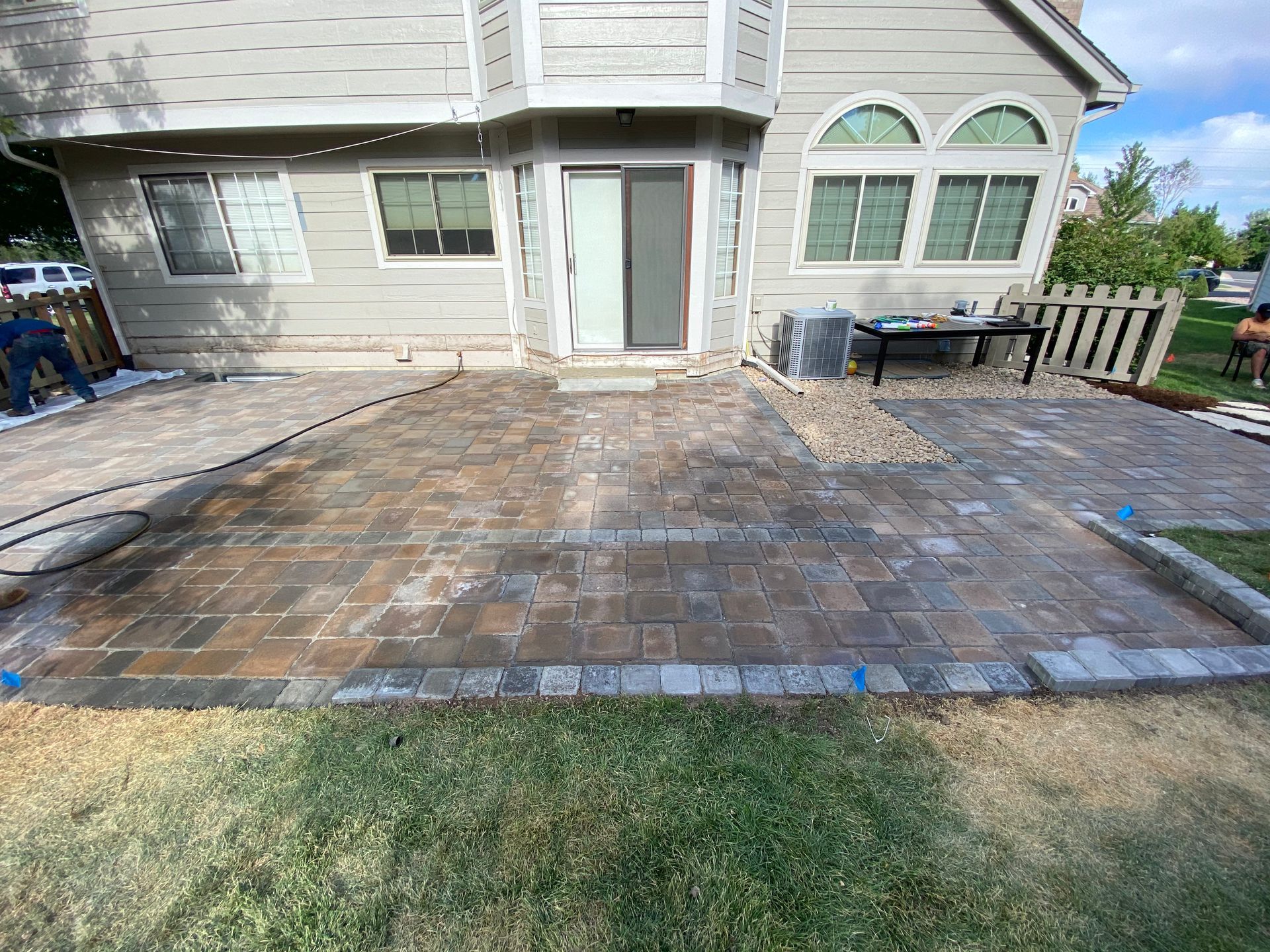 Backyard patio with brick pavers, bordering grass, and a house in the background.