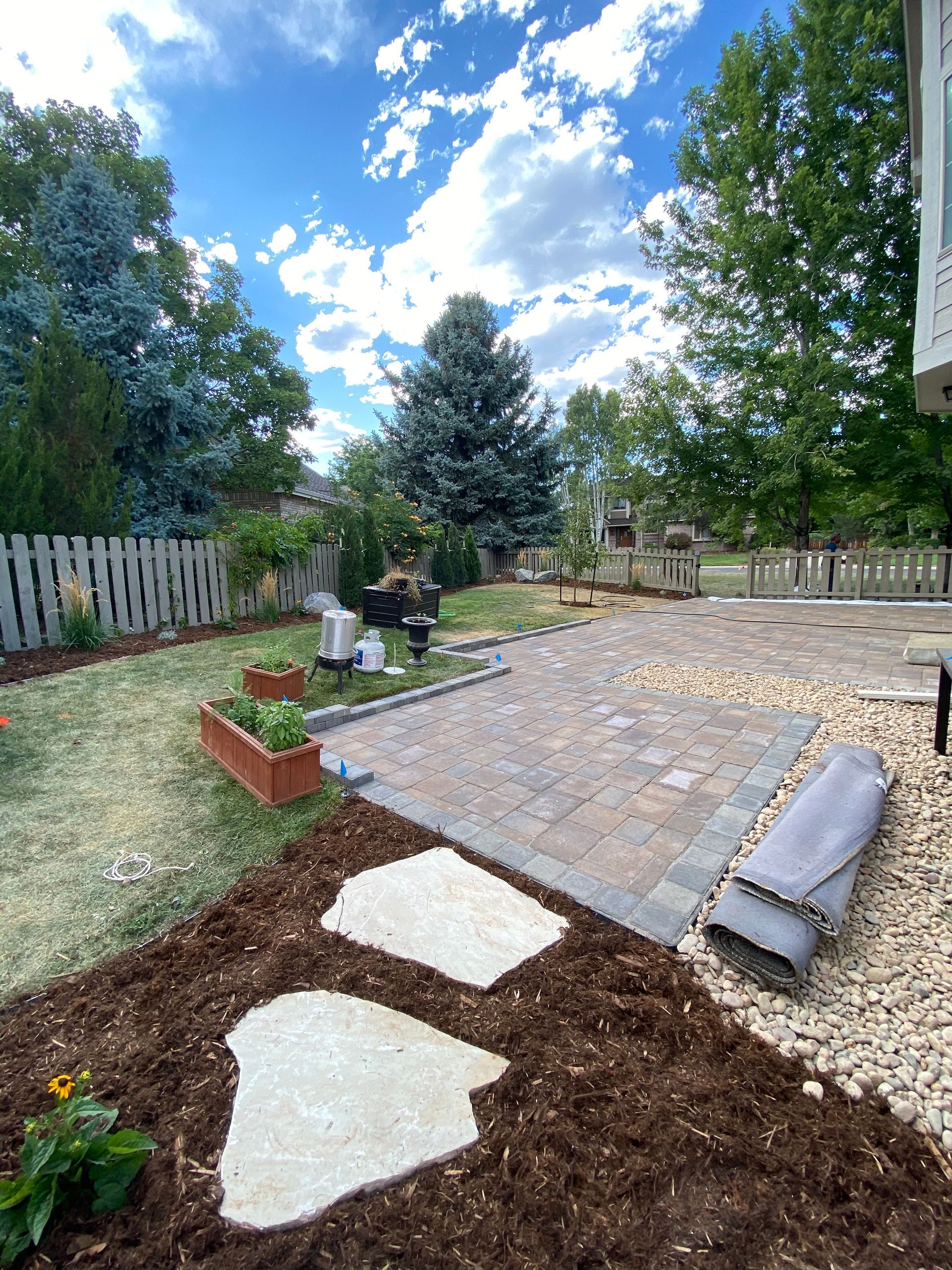 Backyard with stone patio, stepping stones, wooden fence, trees, and blue sky.