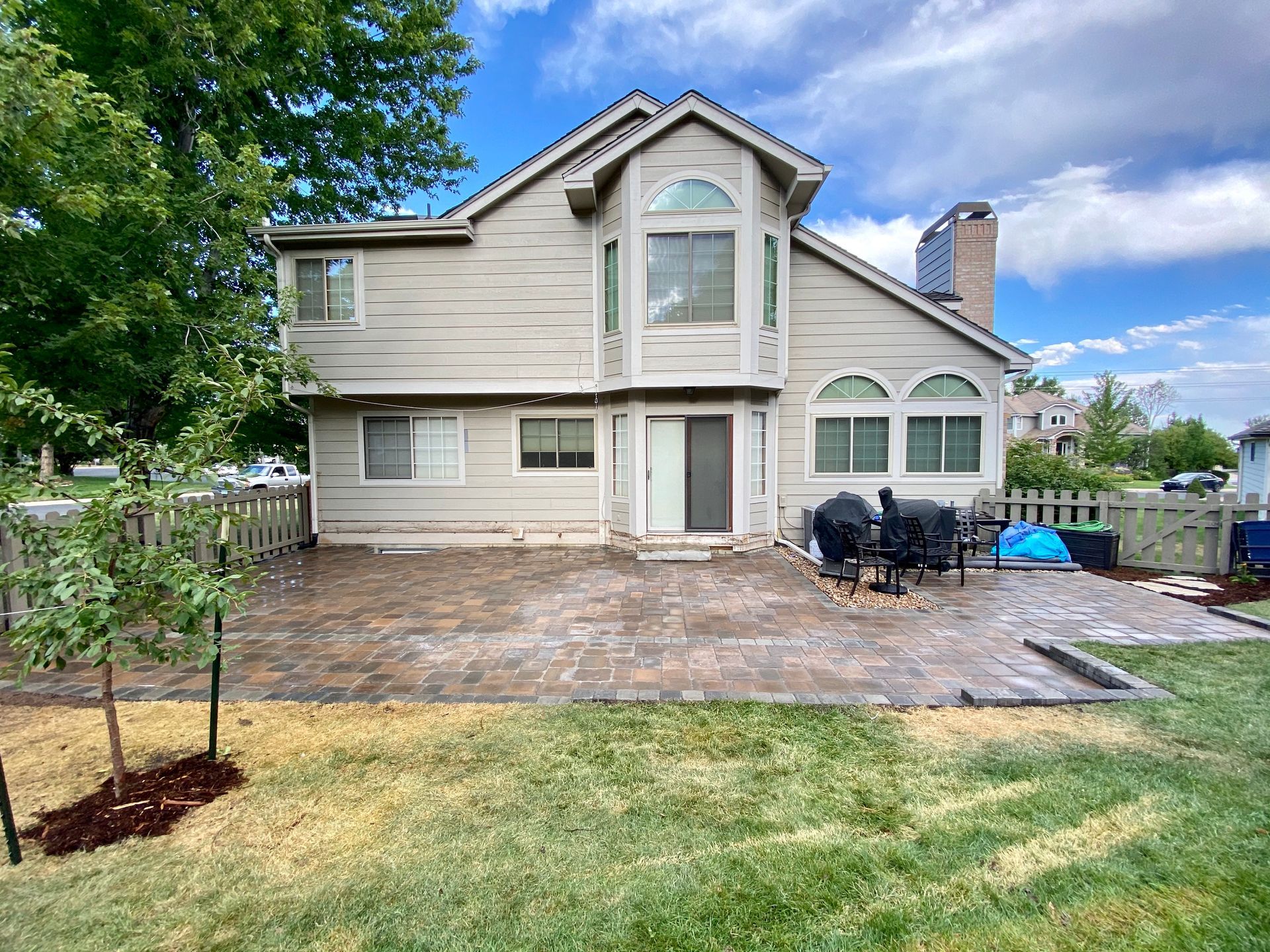Backyard patio with a two-story beige house and a brick patio. Includes trees, lawn, and furniture.