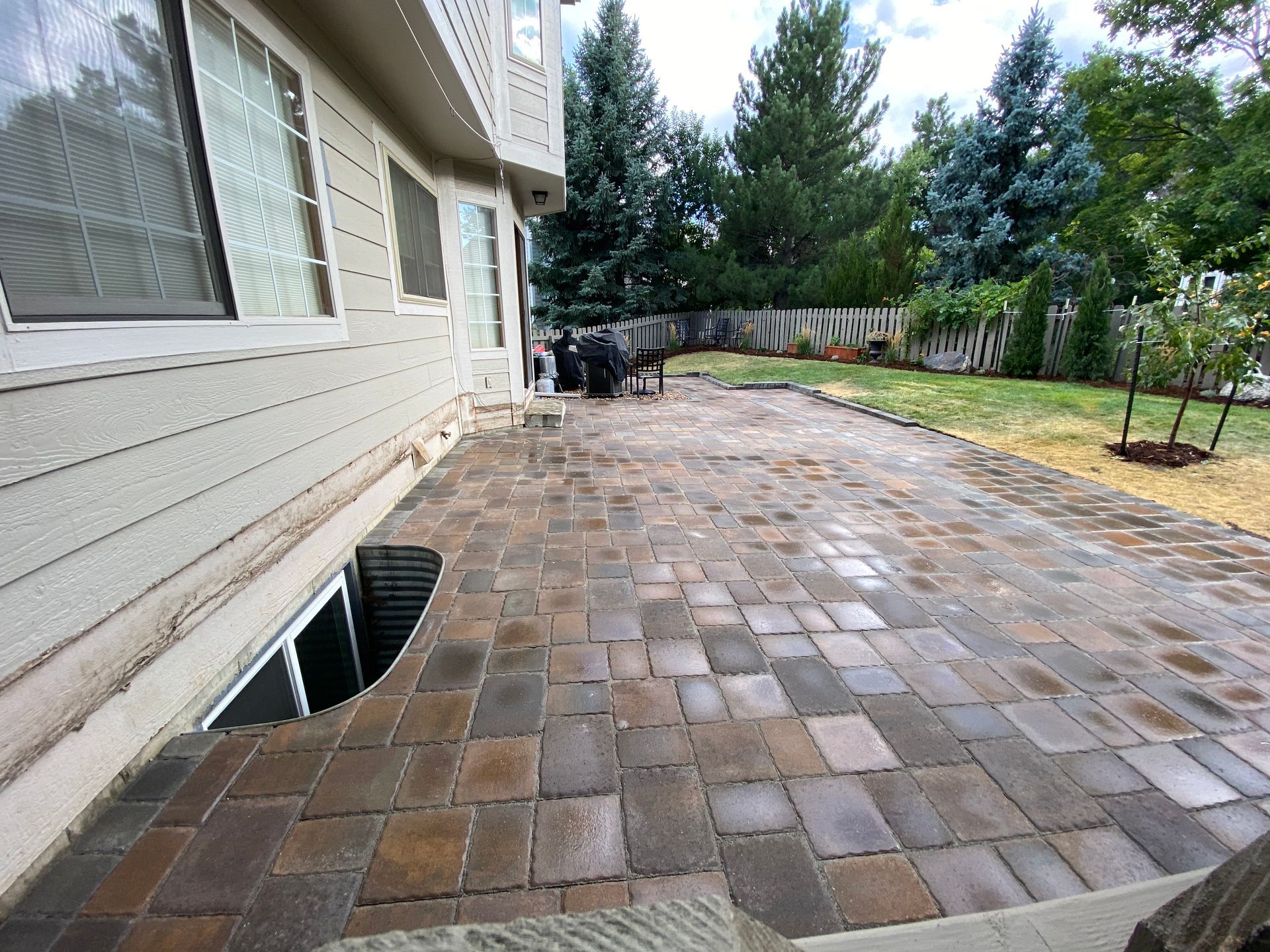 Brick patio next to a house with a window well, trees, and a grassy backyard.