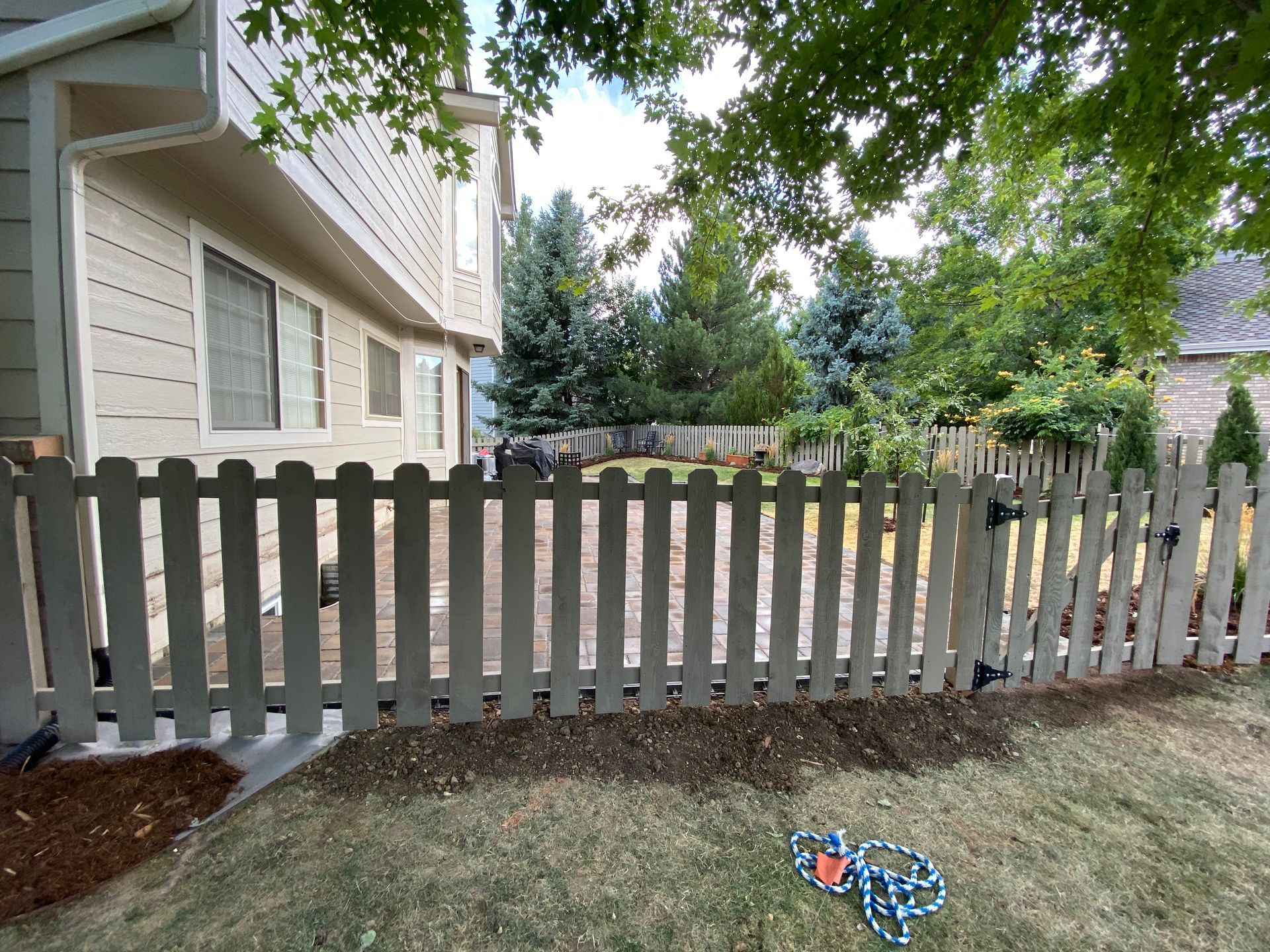 Wooden picket fence in a backyard, with a house and trees in the background.