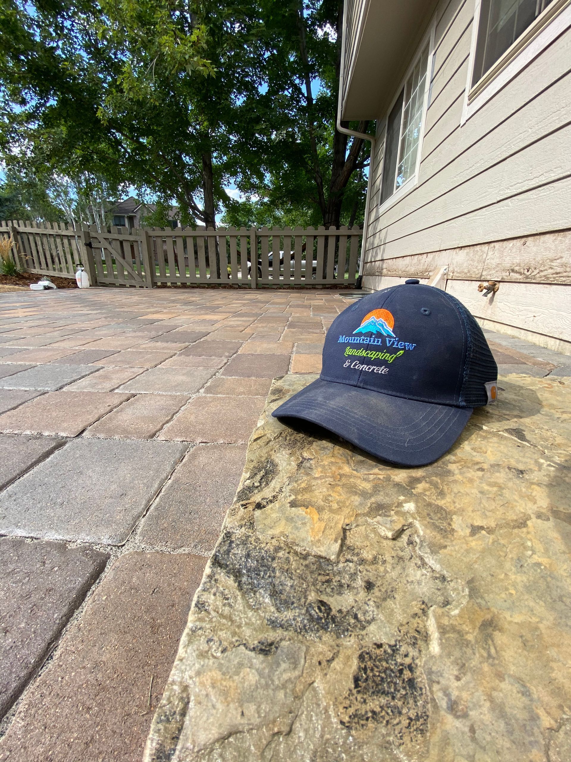 Blue cap with logo rests on stone edge of a patio, near a house.