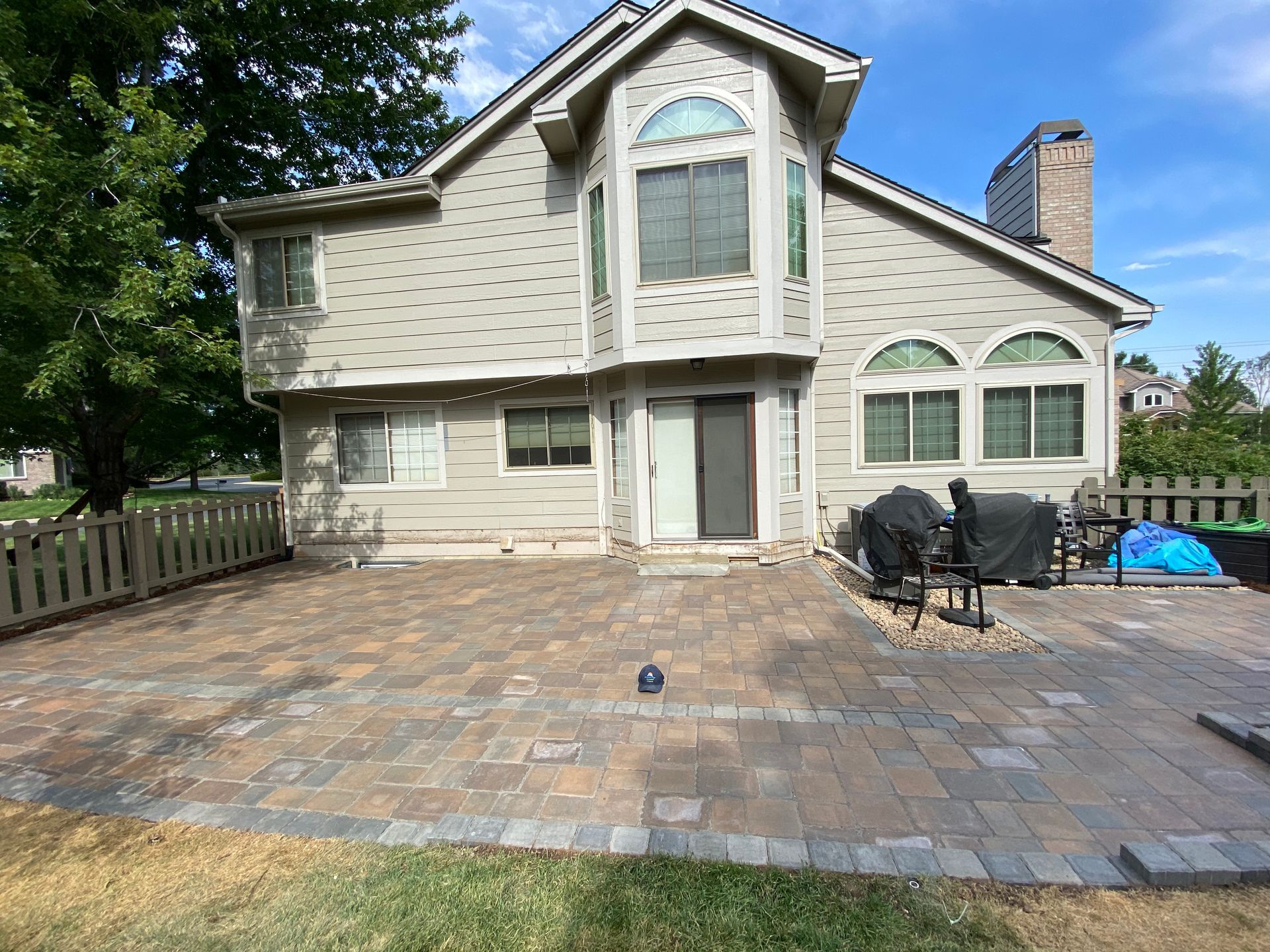 A light beige two-story house with a brick patio, a fence, and outdoor grill on a sunny day.