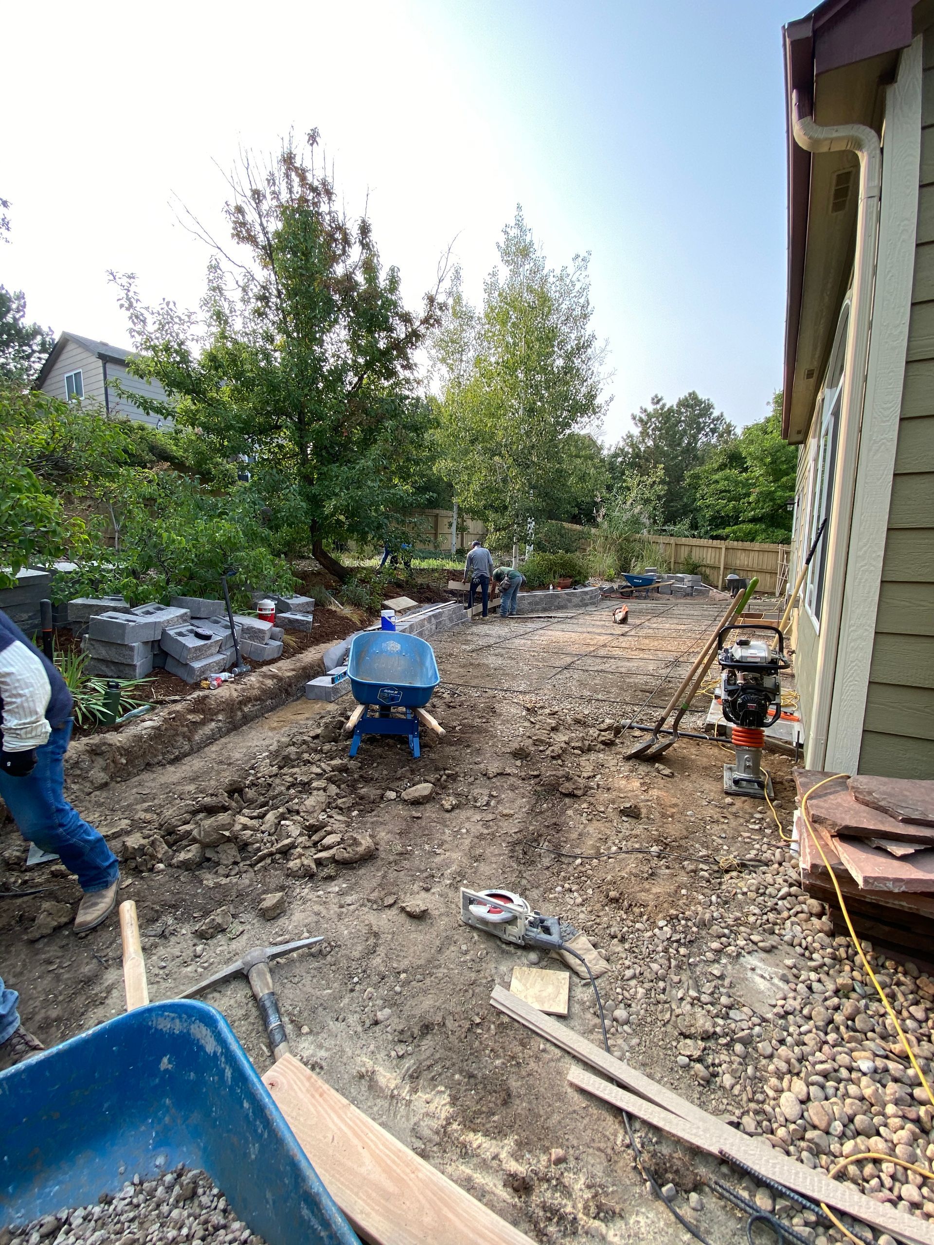 Construction site with workers, wheelbarrows, and tools; gravel and dirt path next to a building.