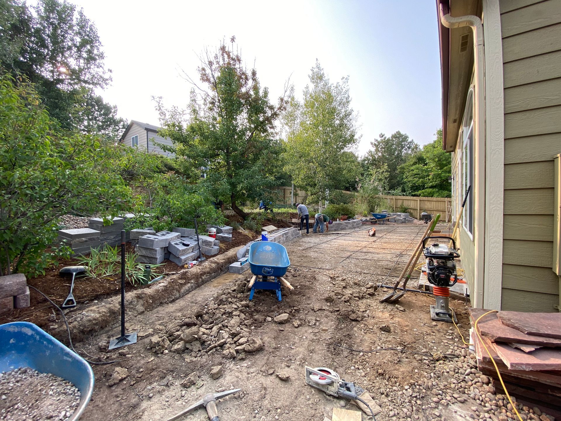 Construction site: workers laying pavers in a backyard. Blue wheelbarrow, compactor, and trees are visible.