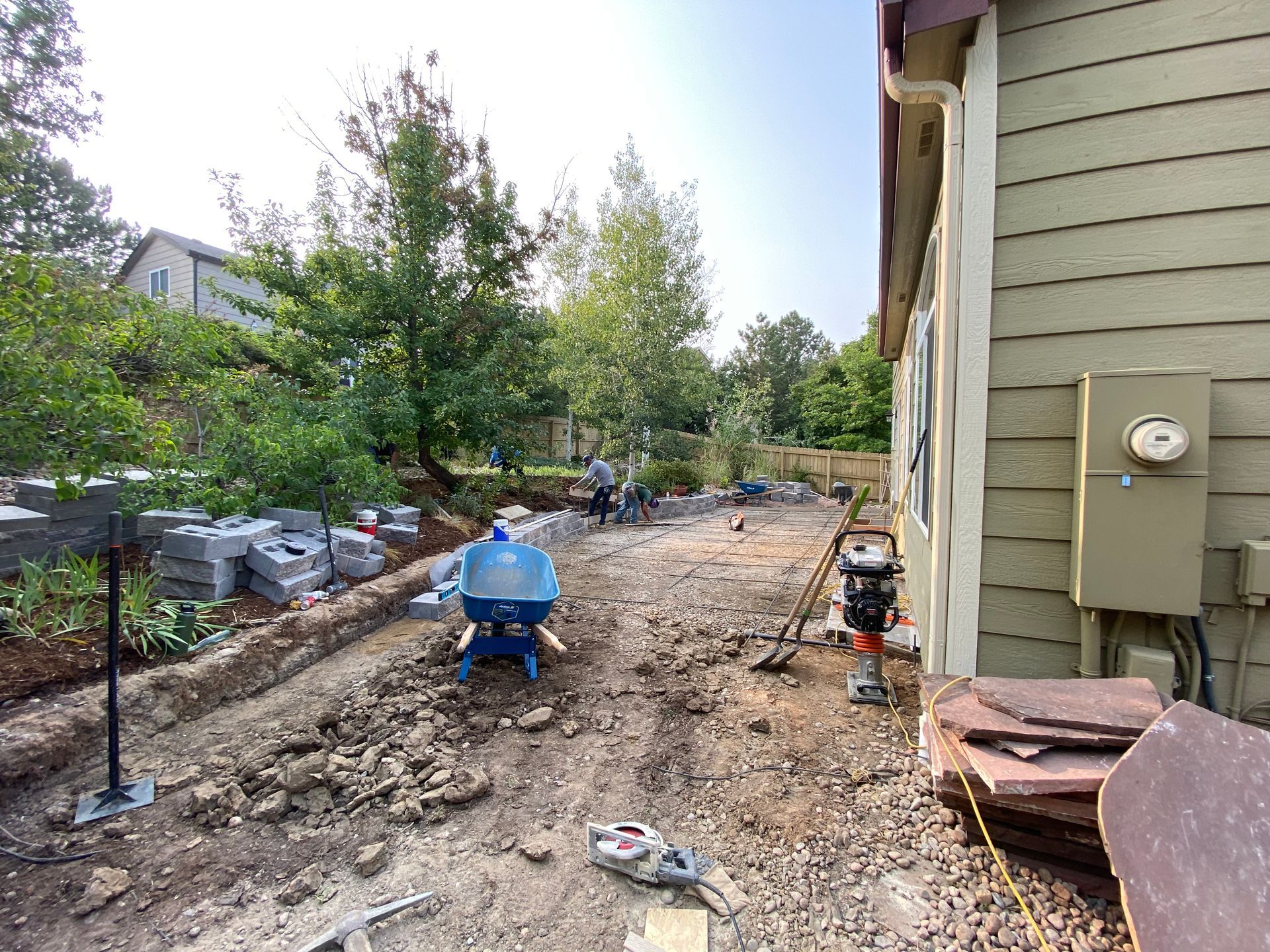 Construction site with exposed dirt, equipment, and workers near a house; blue sky.