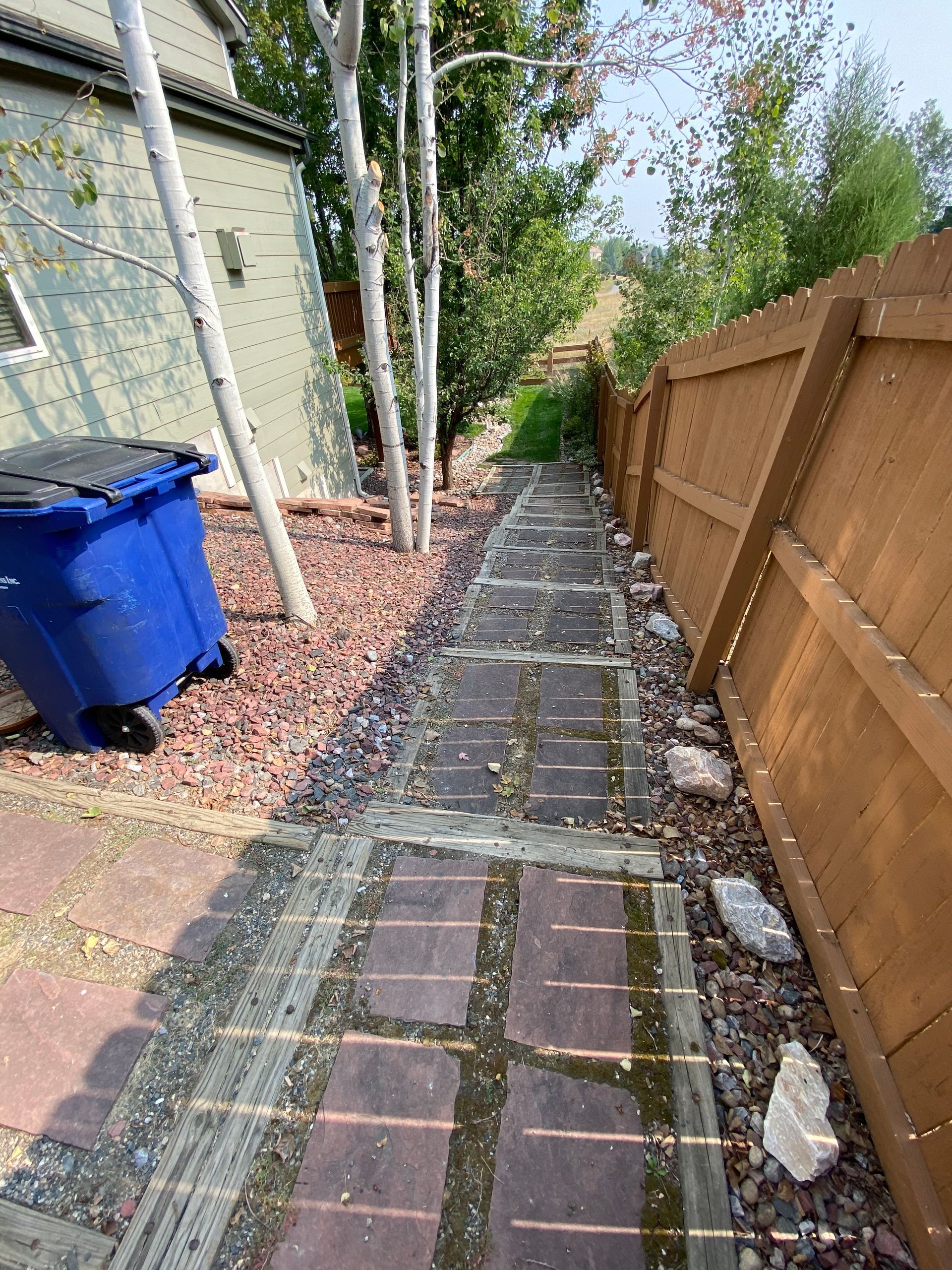 Brick pathway leading uphill between a wooden fence and trees, with a blue trash can to the left.