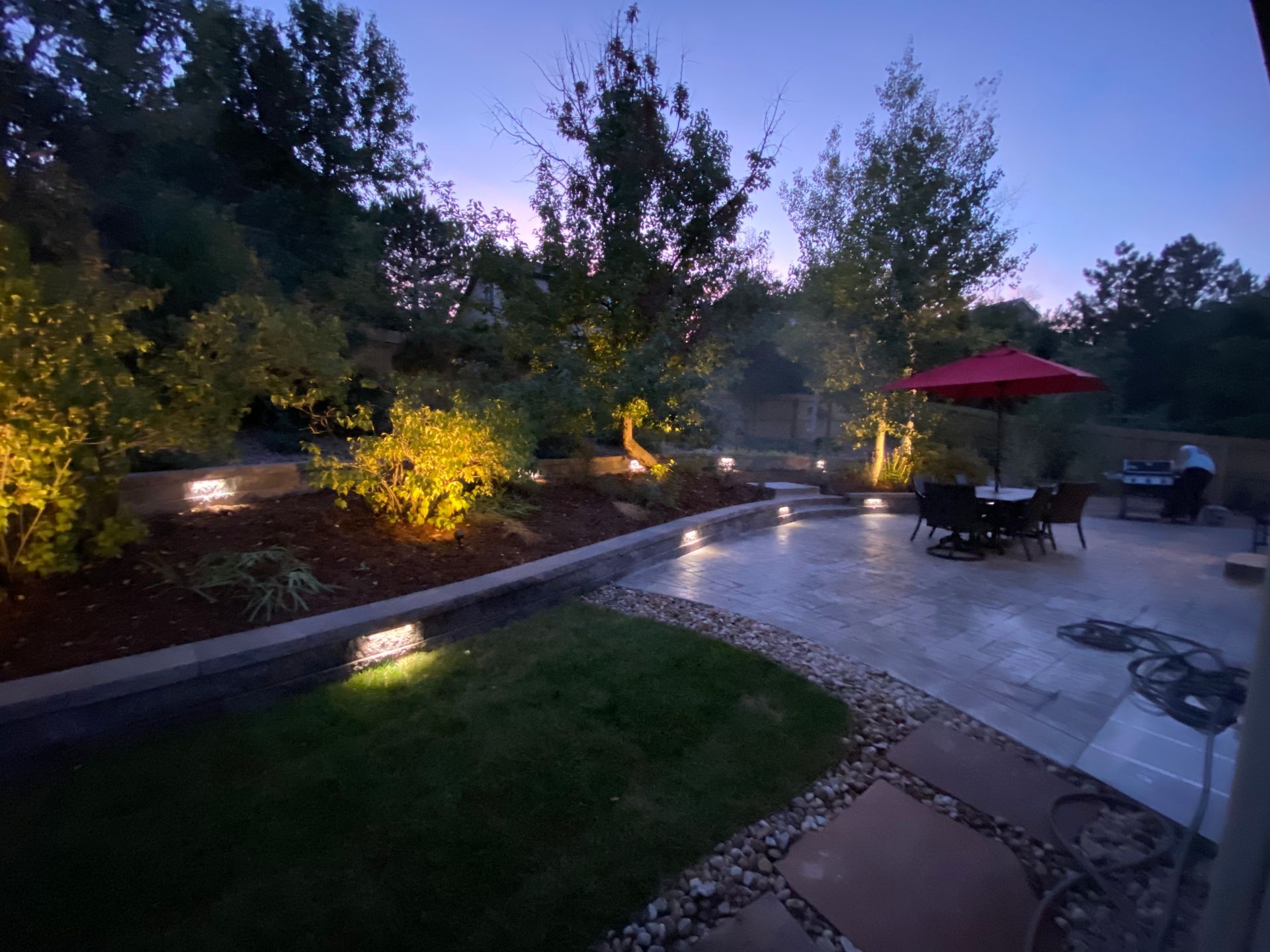 Backyard patio with landscape lighting at dusk. Includes a seating area, lawn, and surrounding trees.
