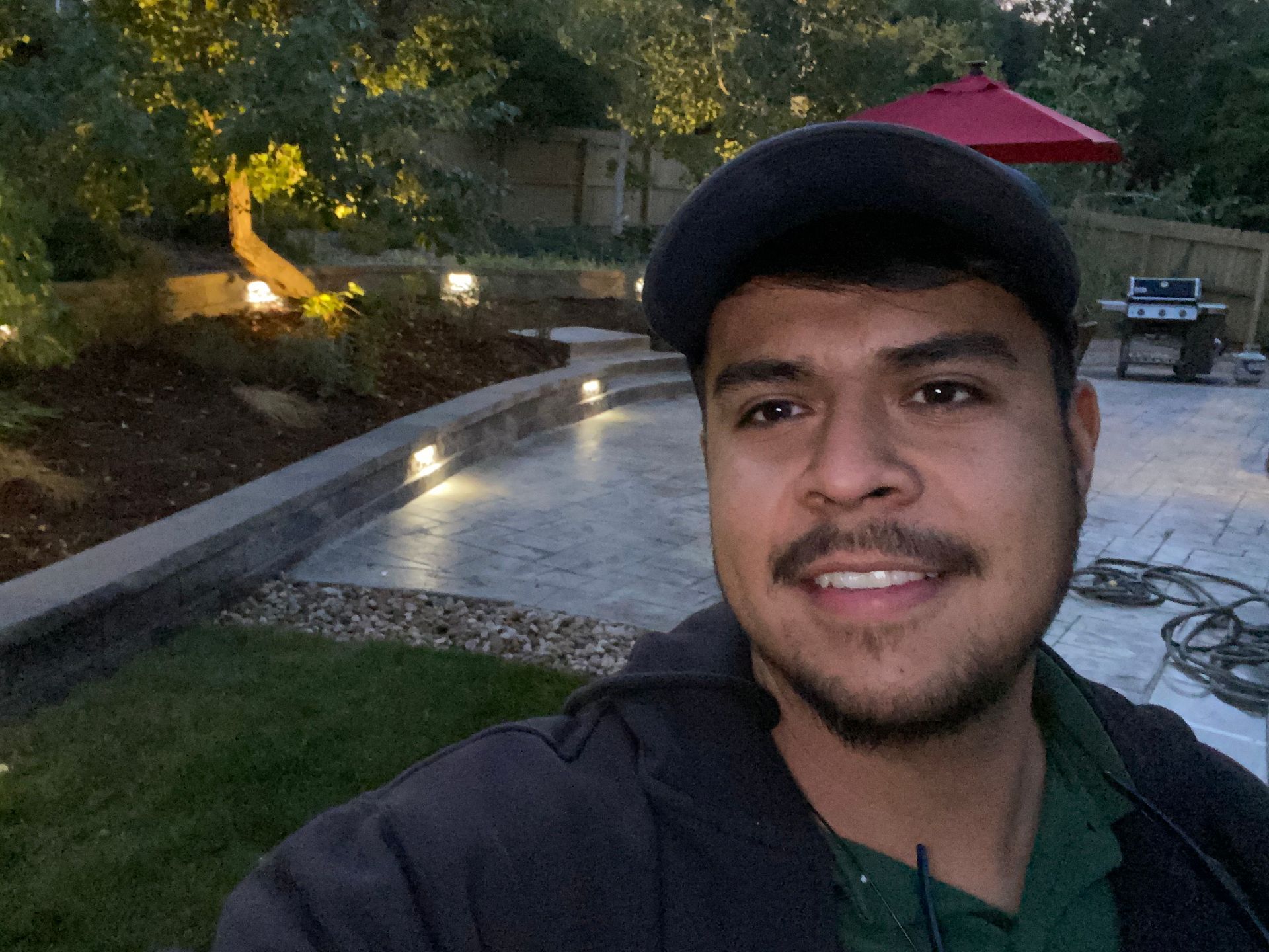 Man with dark cap smiles, standing in a backyard with a lit patio and landscaping.