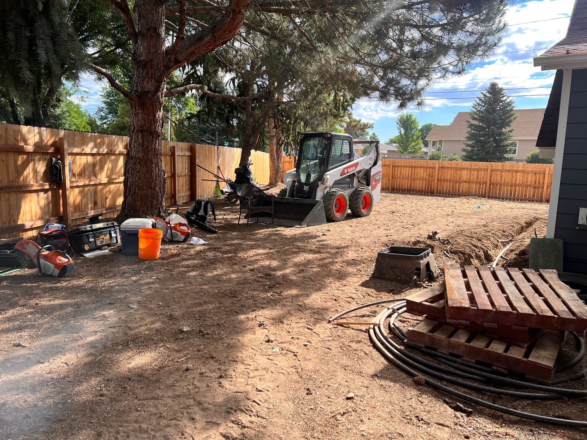 Backyard construction site: Bobcat, tools, dirt, wooden fence, tree, and house.