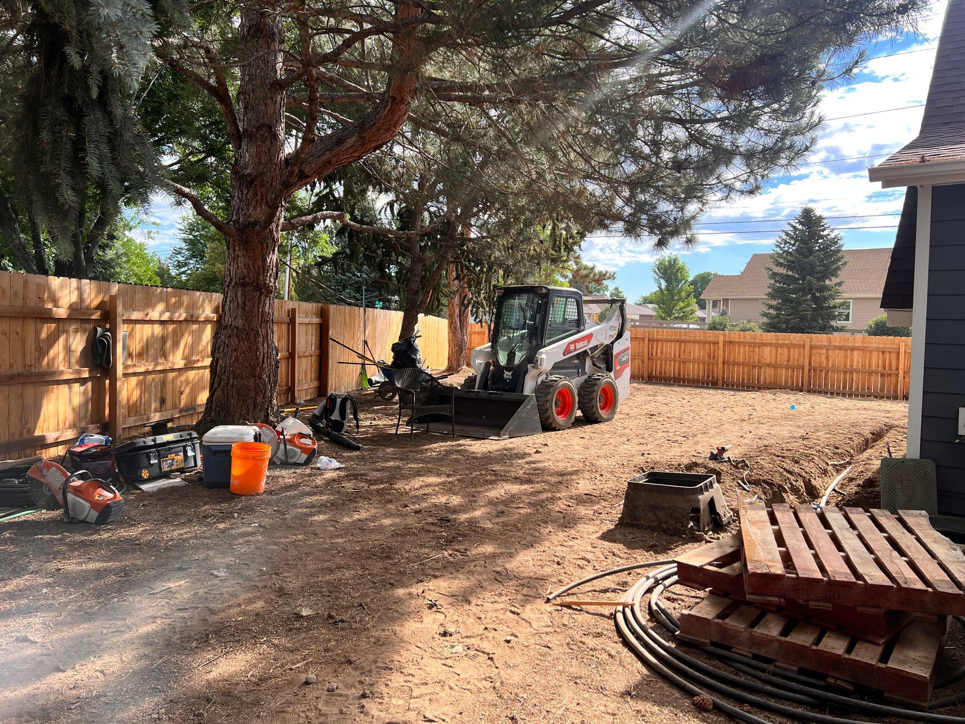 A Bobcat and tools sit in a yard. Construction is in progress near a fence and house.