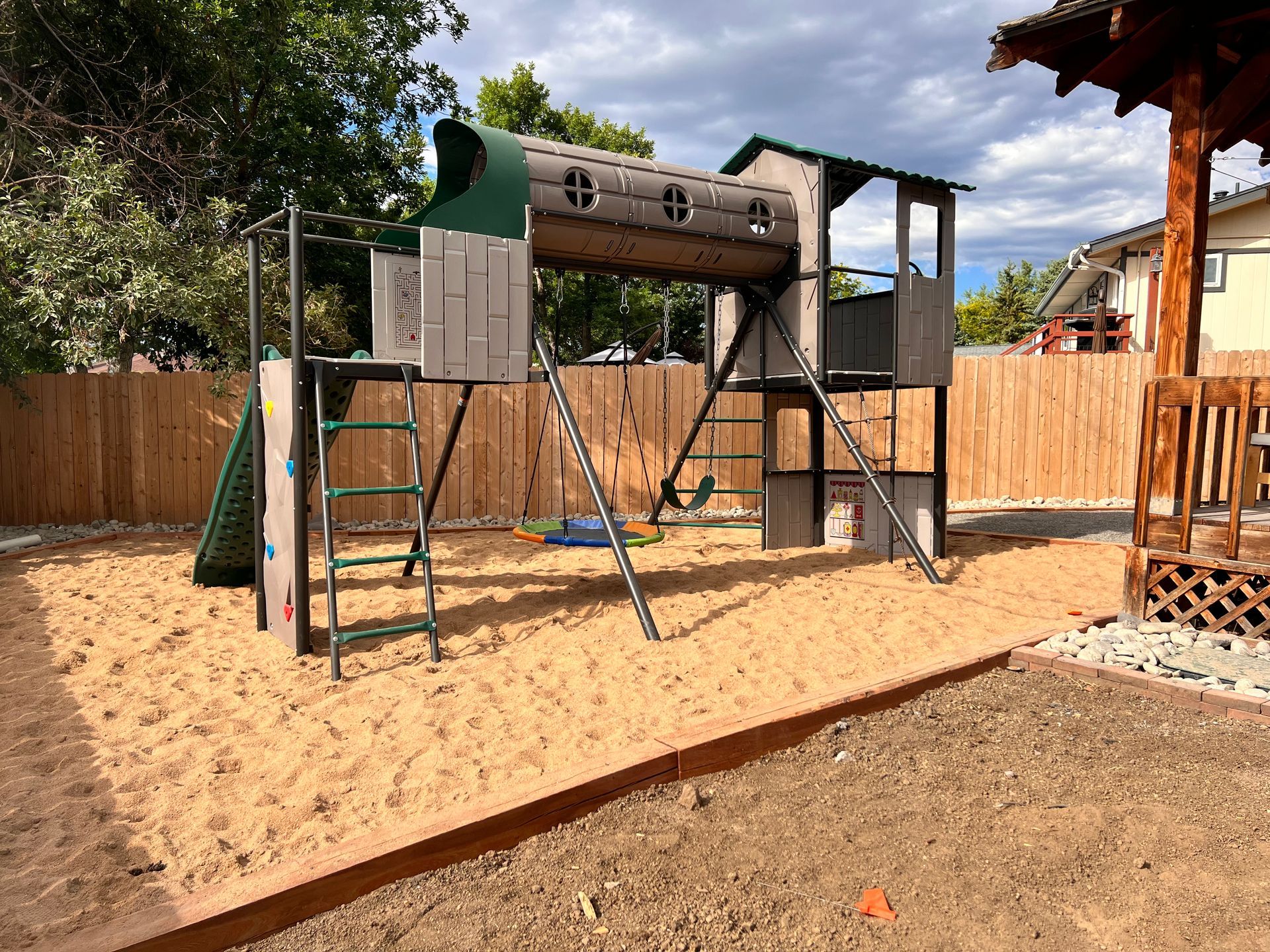 Playground with climbing wall, swings, and wooden structure in a sand pit, fenced yard.