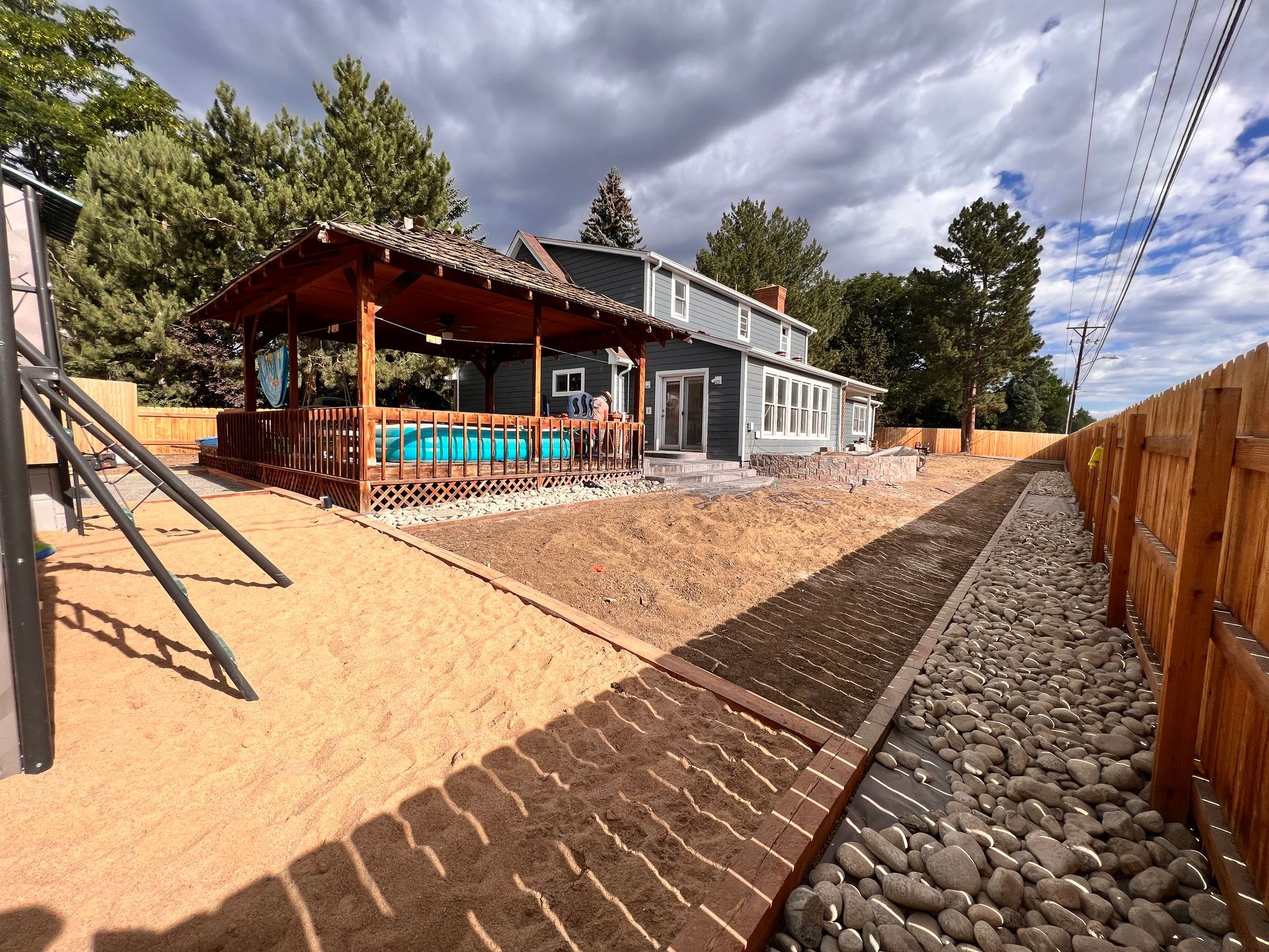 A two-story gray house with a wooden deck and pool, brown gravel and sand pathway, under a cloudy sky.