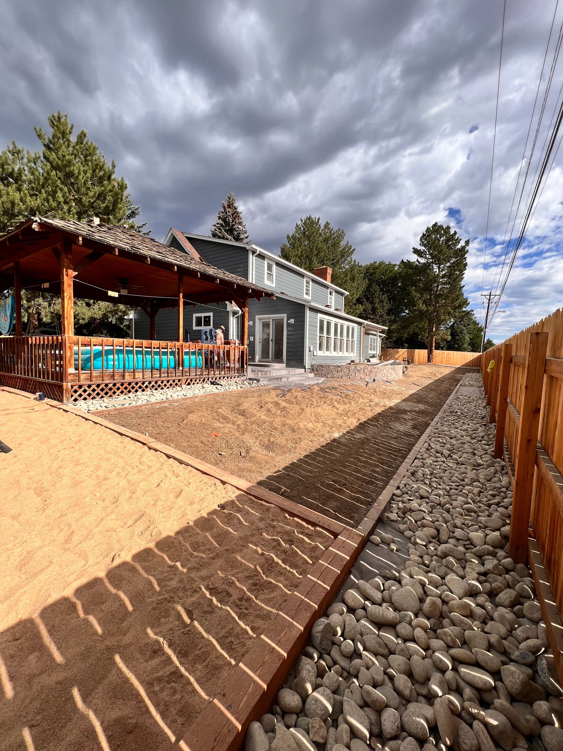 Backyard with pool, wooden deck, gravel path, wooden fence, gray house under cloudy sky.