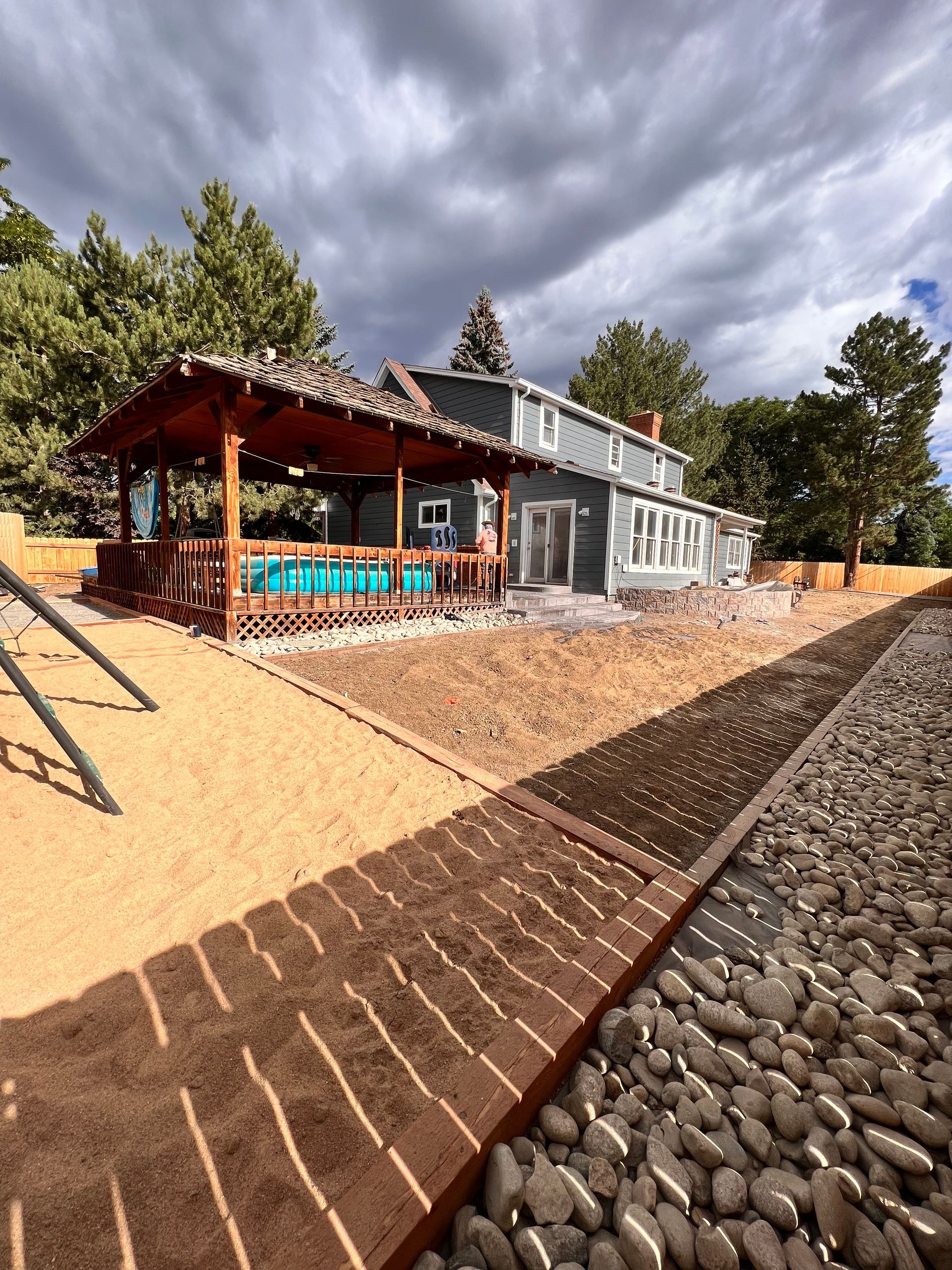 House with a brown deck, tan landscaping, and gray exterior under a cloudy sky.